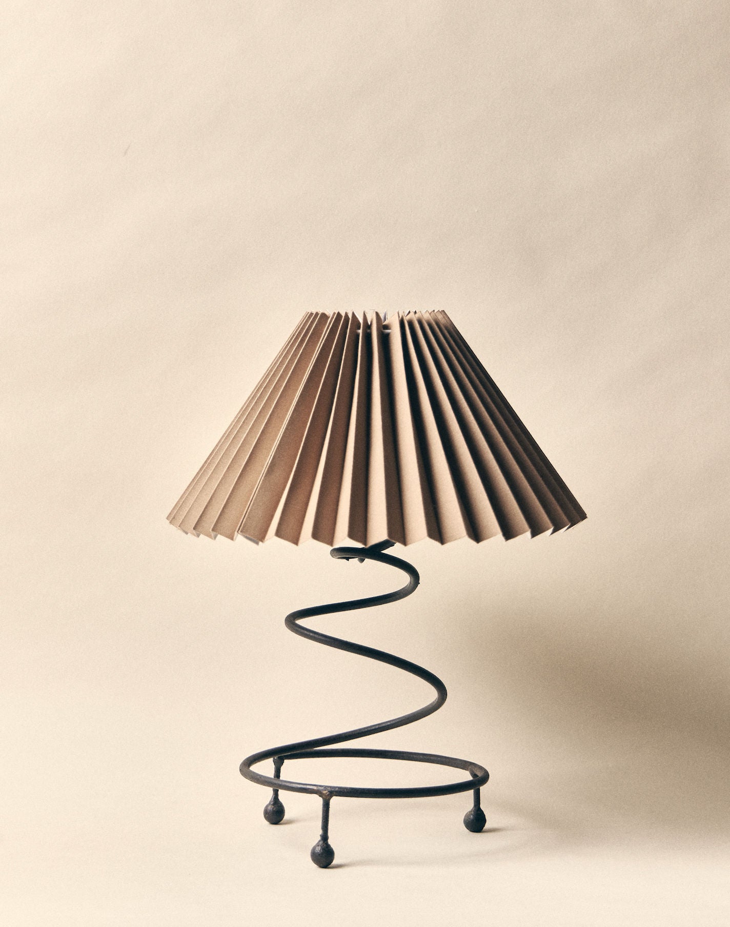 The SÒLER 'Como' Table Lamp features a pleated beige shade and a black wrought-iron spiral base, beautifully set against a matching beige background.
