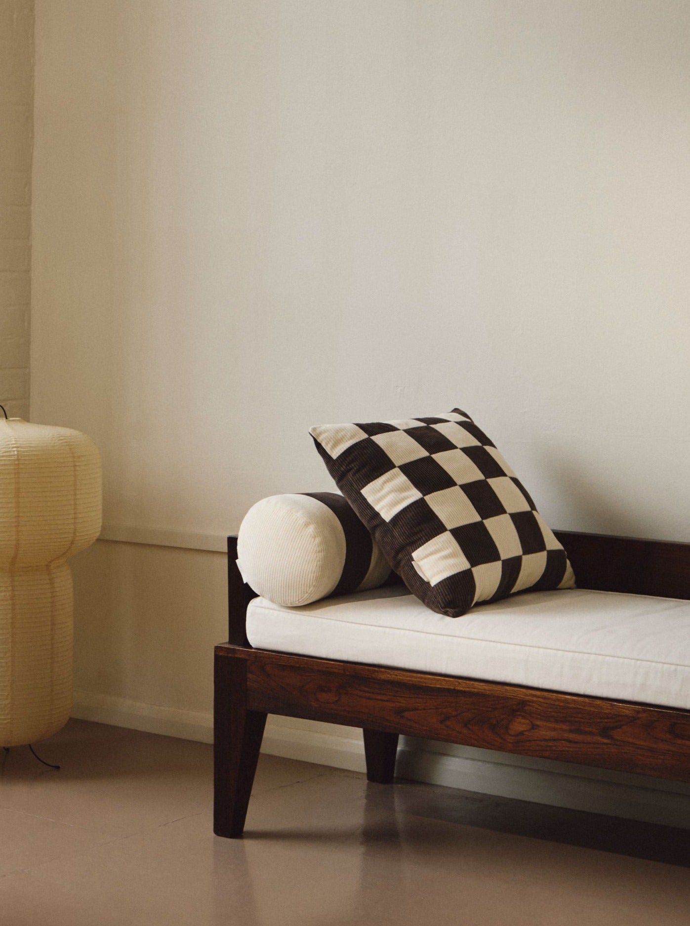 A minimalist room featuring a wooden daybed with white cushions and a vintage-inspired patchwork pillow. Beside it stands a unique, sculptural floor lamp made of thin paper-like material. A small white cube stool, AMÉLIORÉ's Le Carré - Cotton Corduroy - Chalk/Espresso, is also present in the foreground, adding to the room's understated charm.