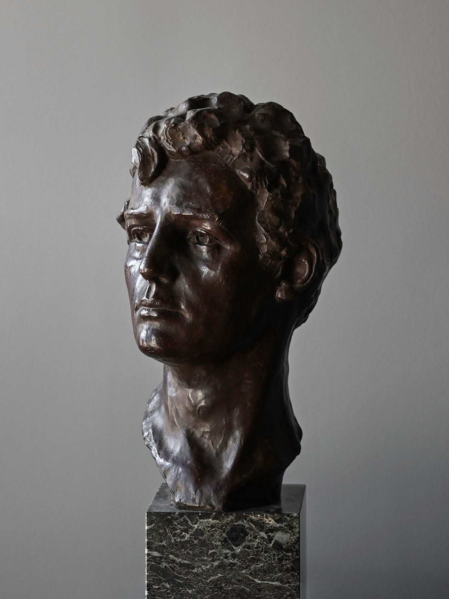 The Bronze Bust of Male by Victor Demanet from Studio OSKLO features wavy hair and lifelike detail, set on a dark marble pedestal and shown against a plain gray background, highlighting Belgian bronze artistry.
