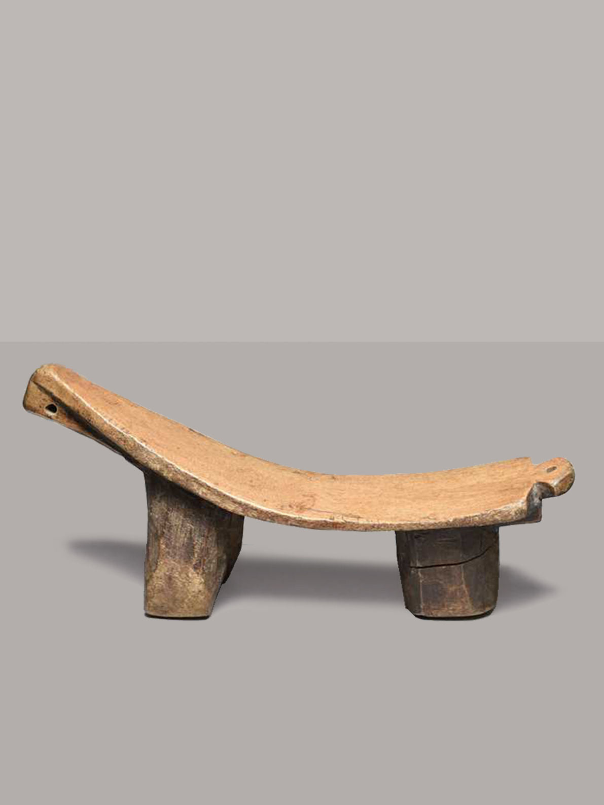 The Lobi Tripod Stool 2 by RELIC LONDON is a traditional wooden headrest with a curved, slightly concave top surface, supported by two solid legs. It is reminiscent of African tripod wooden stools from Burkina Faso. The rustic design and light brown, weathered finish show signs of age and use, set against a plain, neutral gray background.