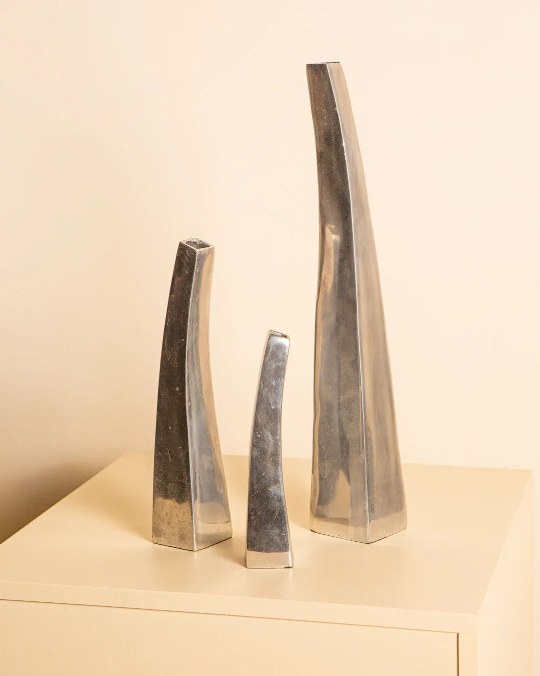 Three silver, elongated abstract sculptures of varying heights stand on a light beige rectangular pedestal against a plain background, exuding timeless elegance reminiscent of the Treaptyque Set of 3 Italian Vases in Cast Aluminum 80's.