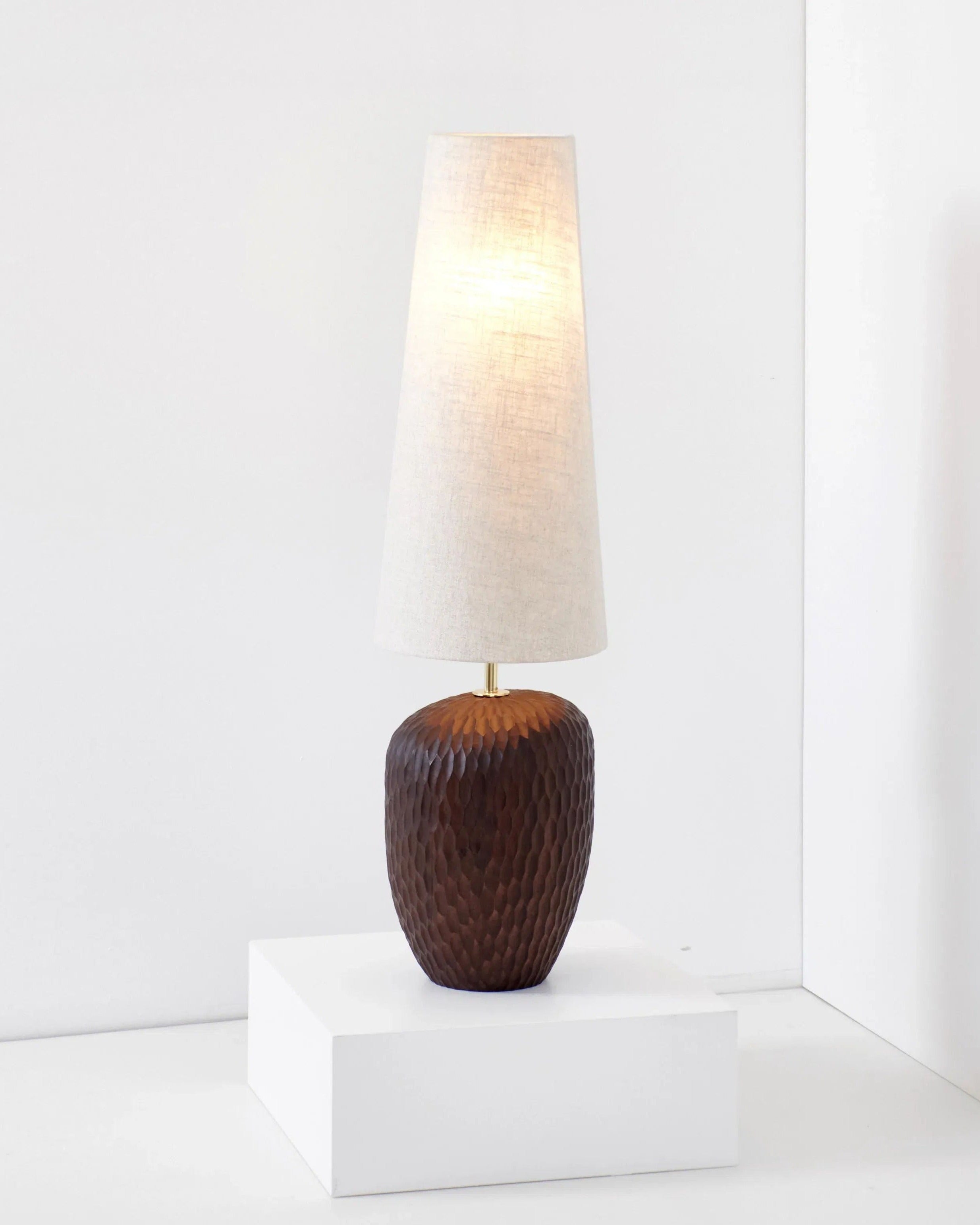 A Large Foz Lamp by Project 213A with a textured, solid wood base and a tall, tapered white lampshade is displayed on a white pedestal. The lamp is set against a minimalist white background.