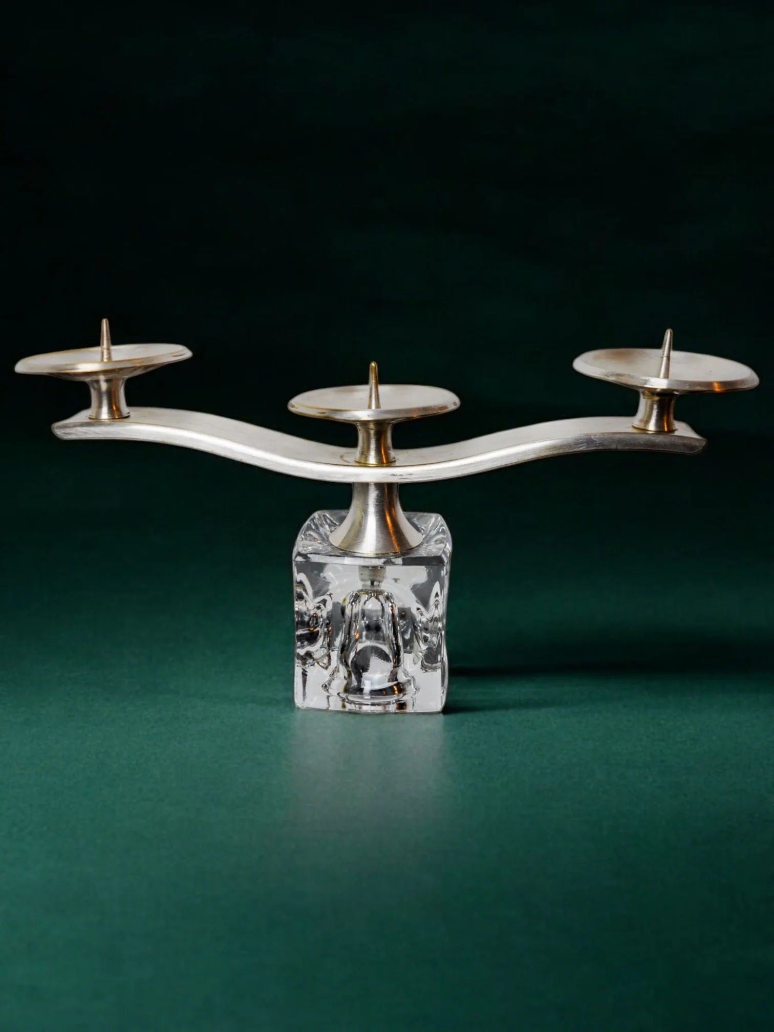 The Ice Cube Candleholder by Peill & Putzler 70s from Malamar Studio, a modern silver three-arm piece with a sculpted clear glass base, is shown on a green surface with a dark green background.