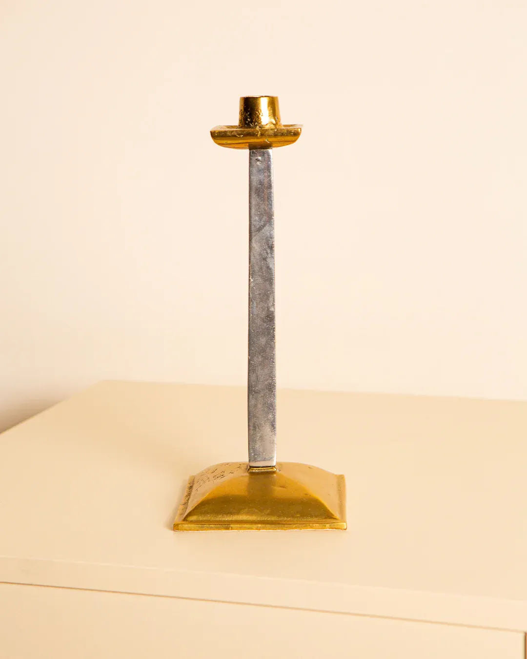 The Brutalist Candleholder by David Marshall 80's from Treaptyque is a tall, slender piece with a square base and gold finish. Its metallic shaft contrasts smoothly with the golden base and top, offering an elegant look on any light surface.
