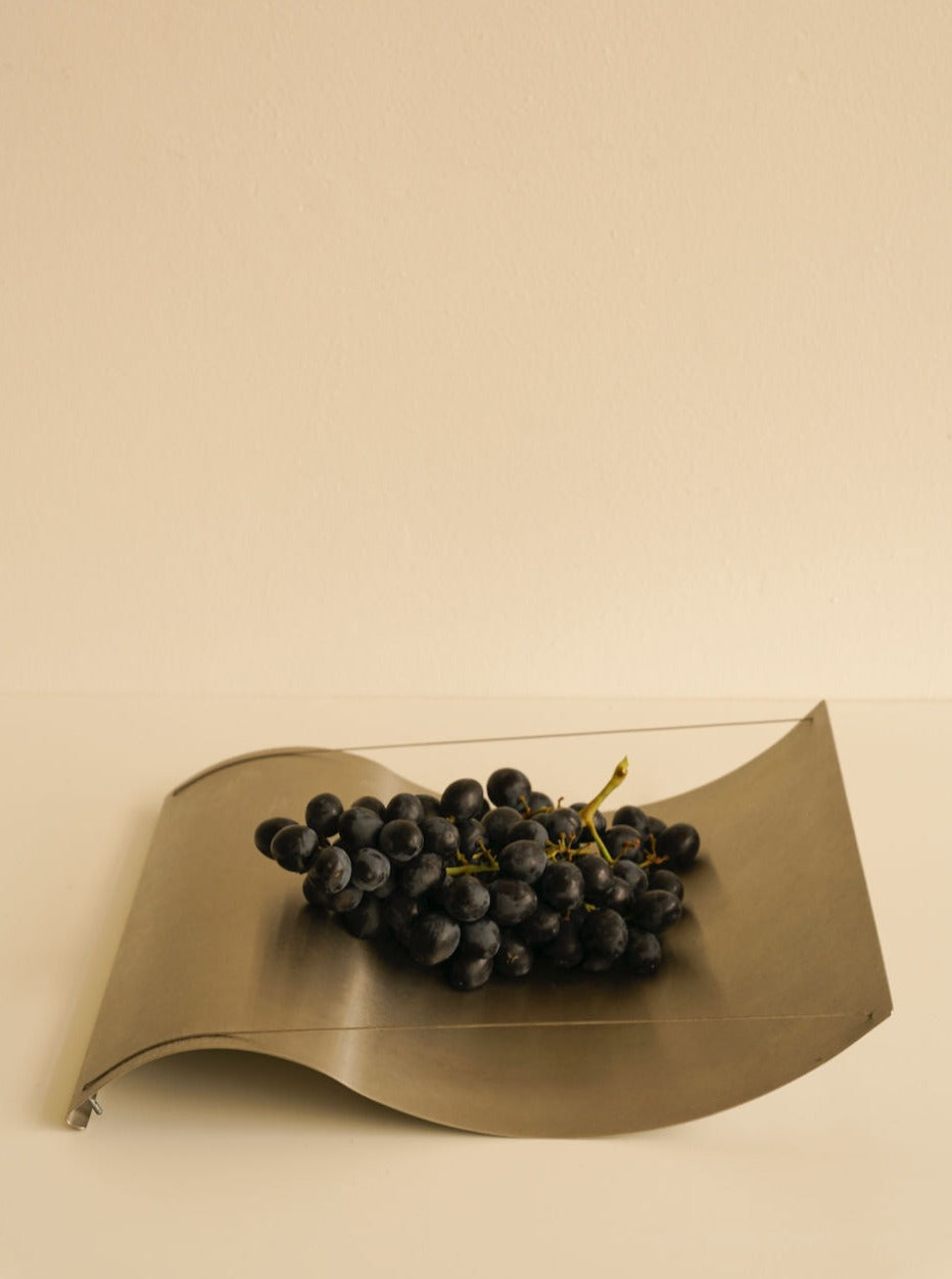 A cluster of dark grapes rests on a Les Objets Amsterdam Studio Eldink holder placed against a light beige background. The smooth lines of the holder accentuate the natural shape of the grapes.