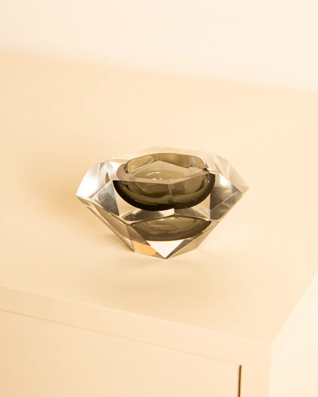 The "Diamant" Vide-poches by Treaptyque, designed in the style of Murano glass art by Flavio Poli in the 80s, features a diamond-shaped, faceted design with a dark interior and rests on a light surface with a soft beige background.