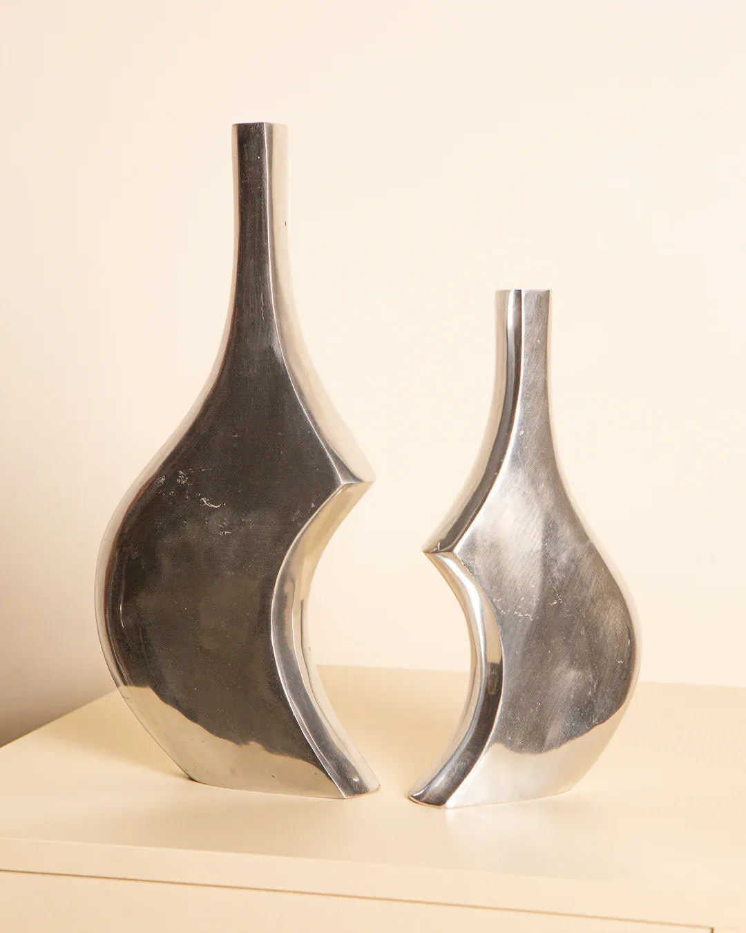 The vintage Pair of 80's Italian Aluminum Vases by Treaptyque, with sleek and modern abstract designs, feature smooth curves. The taller vase sits on the left and the shorter on the right, poised elegantly on a light surface against a neutral background, embodying Italian elegance.