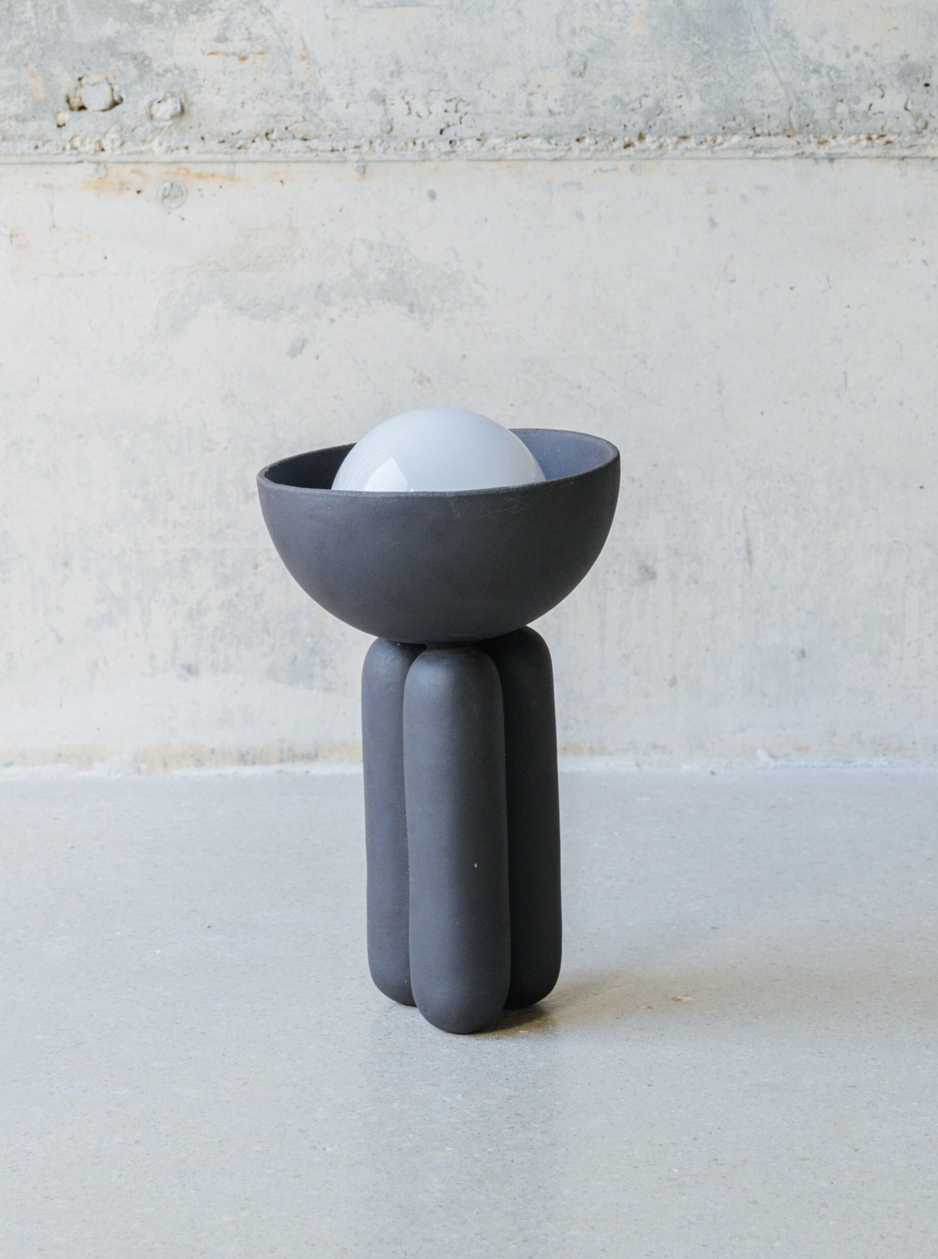 Introducing the Table Lamp Half-sphere M MOOR by Lisa Allegra, a modern and abstract sculpture with a dark, round base supported by four cylindrical legs. It features a semi-spherical bowl that cradles a smooth, white sphere which serves as sculptural lighting. This piece is beautifully accentuated against a minimalistic, unfinished concrete background.