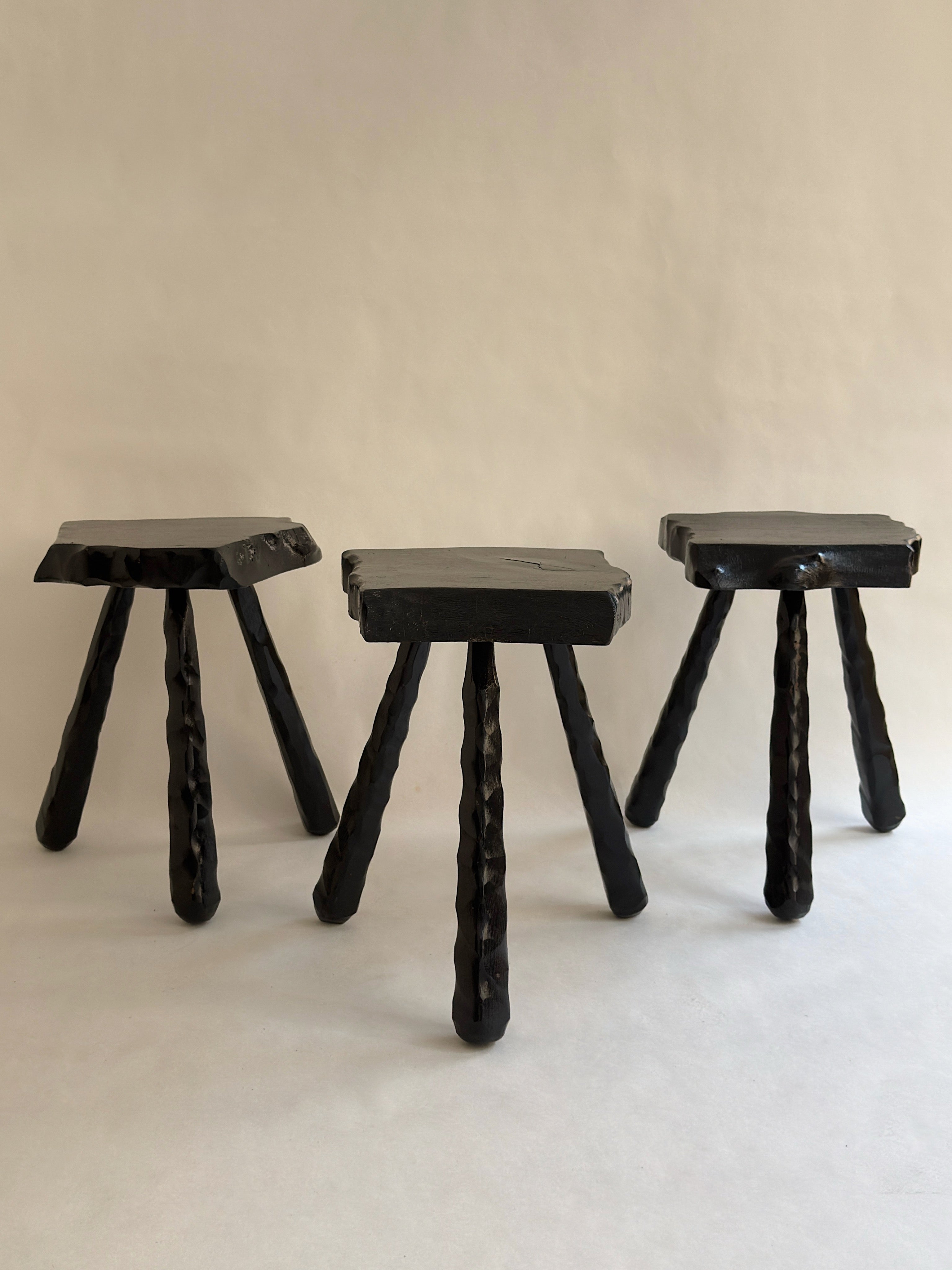 The Trio of Brutalist Stools by Galerie Storm features three handcrafted black wood stools with square seats and textured cylindrical legs, showcasing a rustic, mid-century brutalist style on a plain light background.