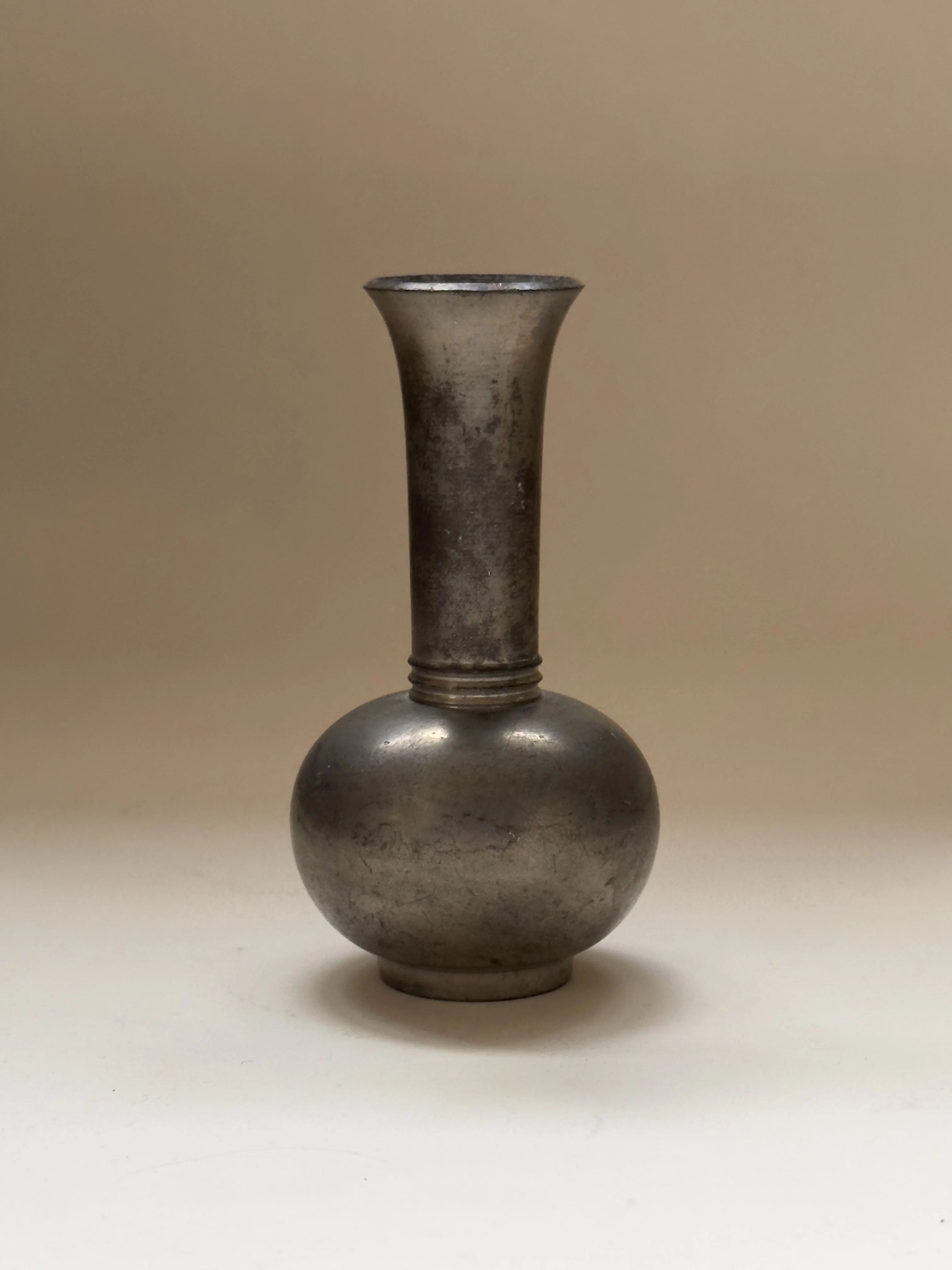 A tall, slender Just Andersen Pewter Vase by Galerie Storm with a narrow neck and round base is set against a simple beige background. Its slightly worn, matte surface exudes vintage charm reminiscent of classic styles.
