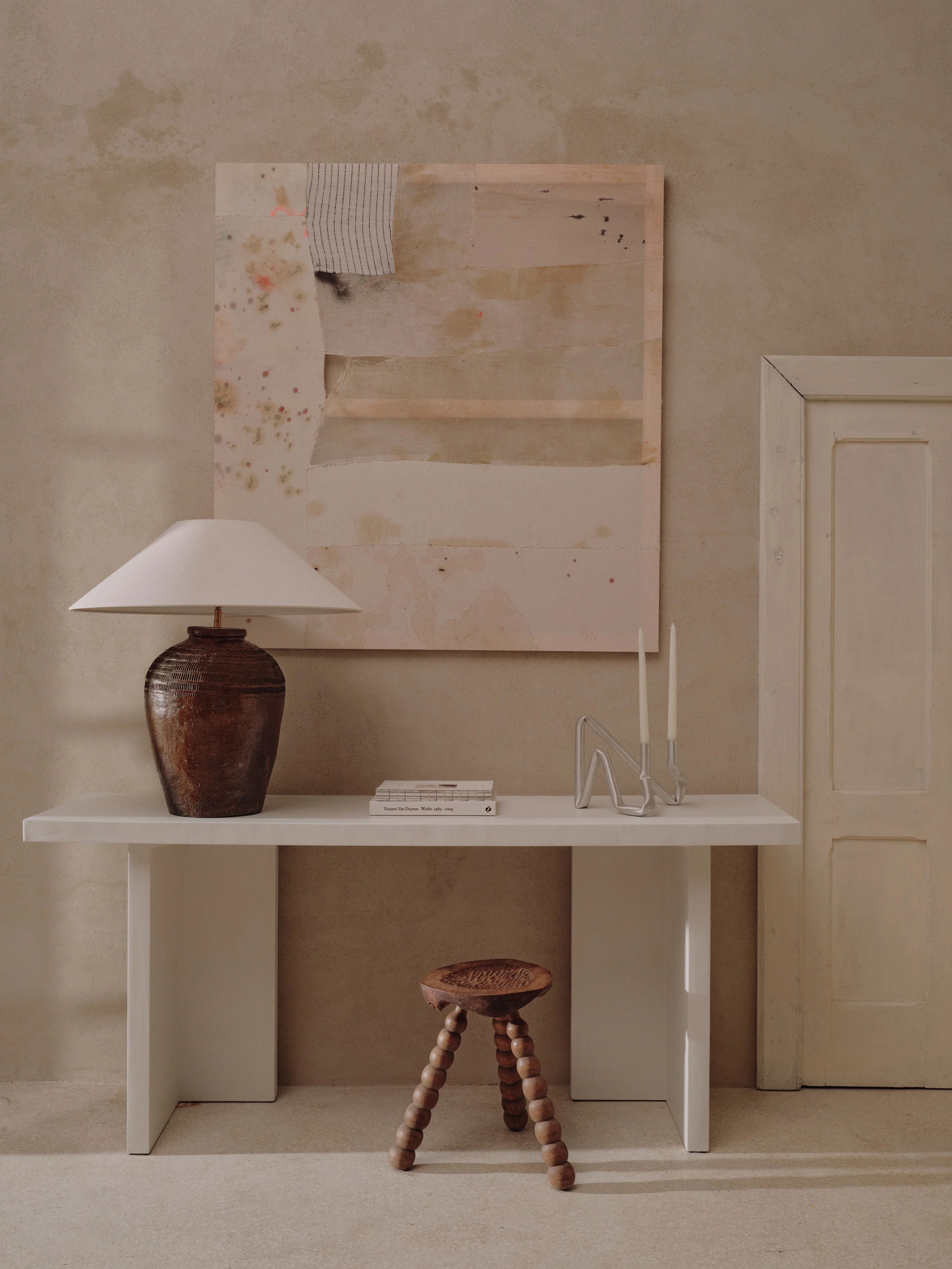 A minimalist room featuring the "Niccolò" Console Table by MarlotBaus, designed with artisan craftsmanship, set against a beige wall. The table is adorned with a ceramic table lamp, books, abstract art, and silver candle holders. In front of it sits a wooden stool with round legs, while a closed white door is on the right.