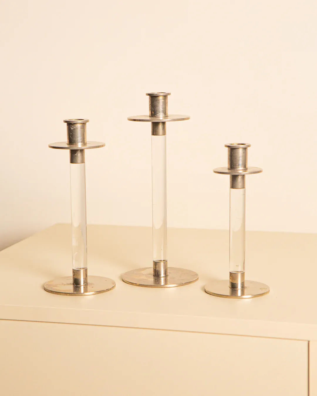 The Treaptyque Set of 3 80's Candleholders features varying height silver candlesticks with minimalist circular bases and cylindrical stems, evoking a retro charm. Displayed on a light surface against a neutral background, they offer a timeless lucite silver blend.