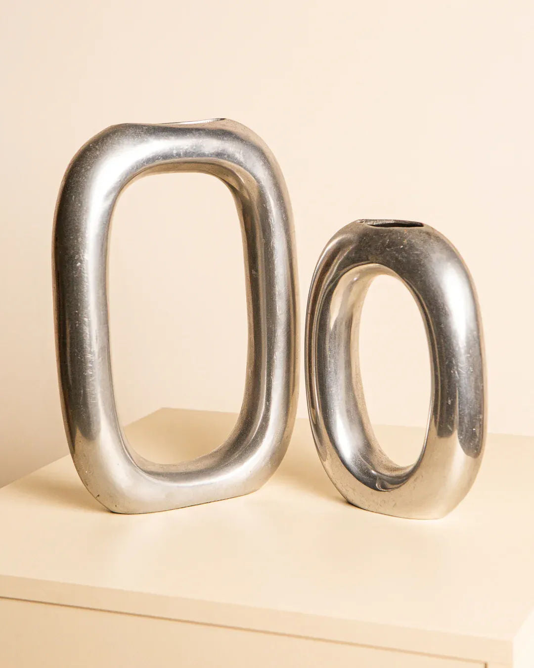 The Pair of Italian Aluminum Vases '80s by Treaptyque exhibit a minimalist design with metallic surfaces and oval openings. Displayed on a cream-colored surface, the taller vase features a more rectangular shape, while its smaller counterpart is slightly rounder. Both vases embody subtle Italian elegance with their modern, shiny appearance.