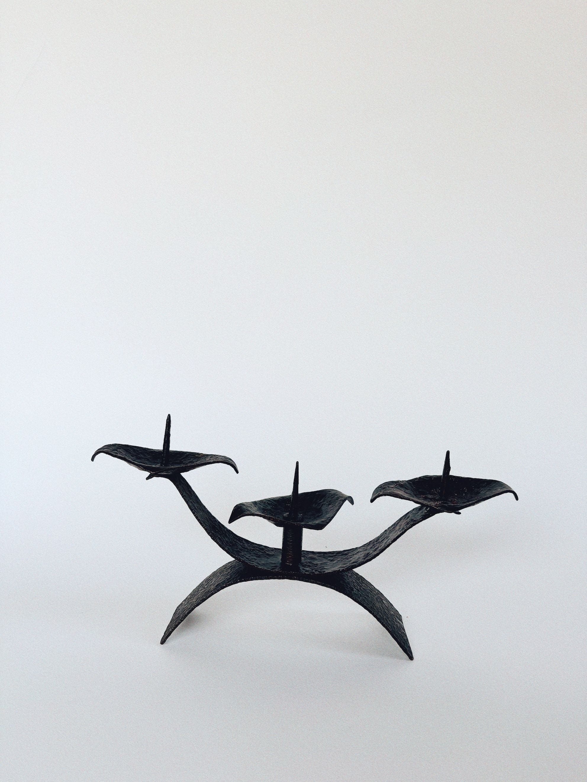 The Huerto 3 Arm Metal Candleholder, designed with sleek, curved lines in minimalist black metalwork, features three prongs for individual candles against a plain white background. It's artisan-made with a modern style.
