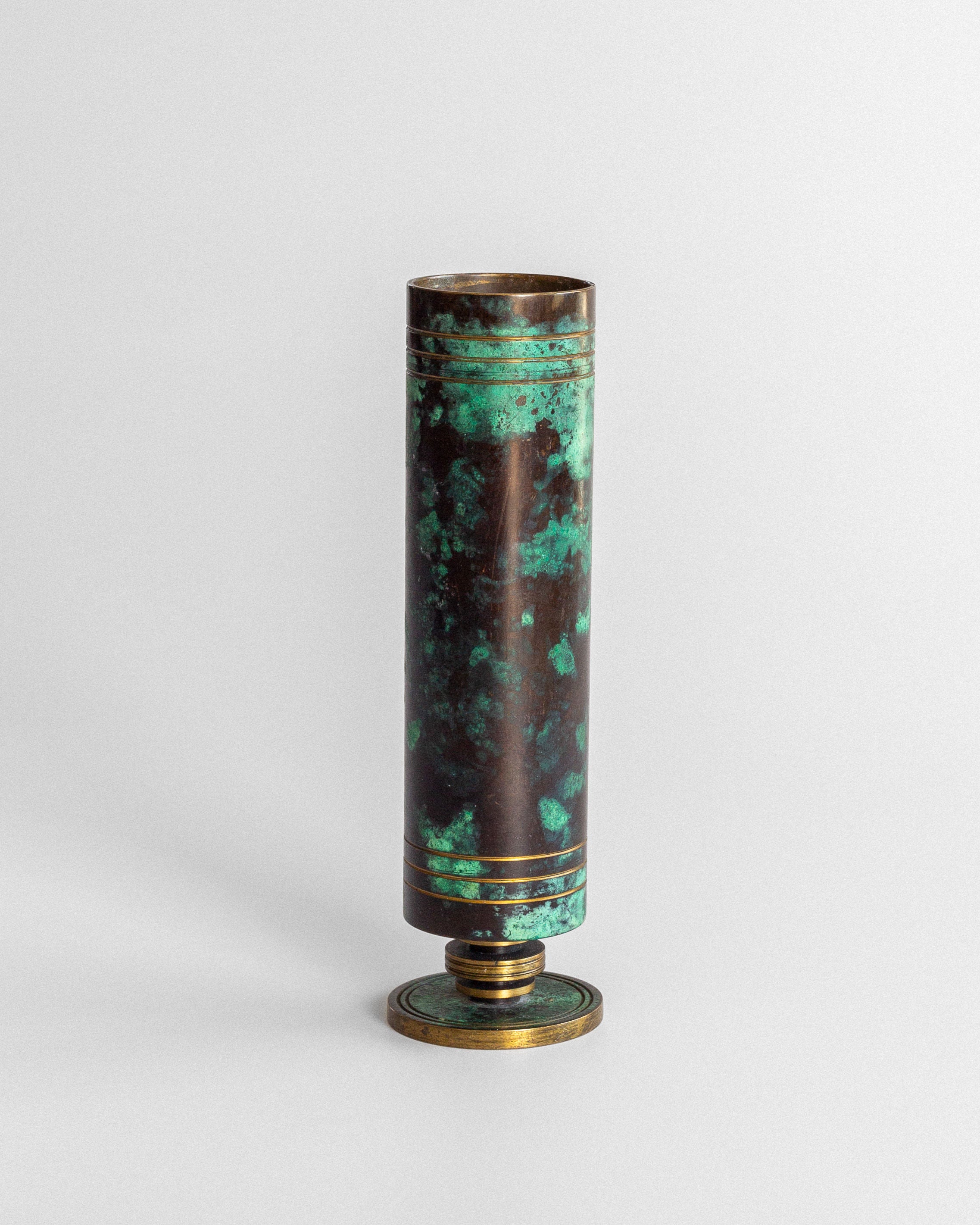 The Spigel Art Deco Bronze and Brass Vase is tall and cylindrical, with a bronze base and rim, displaying emerald green and brown mottled patina, set against a plain light gray background.