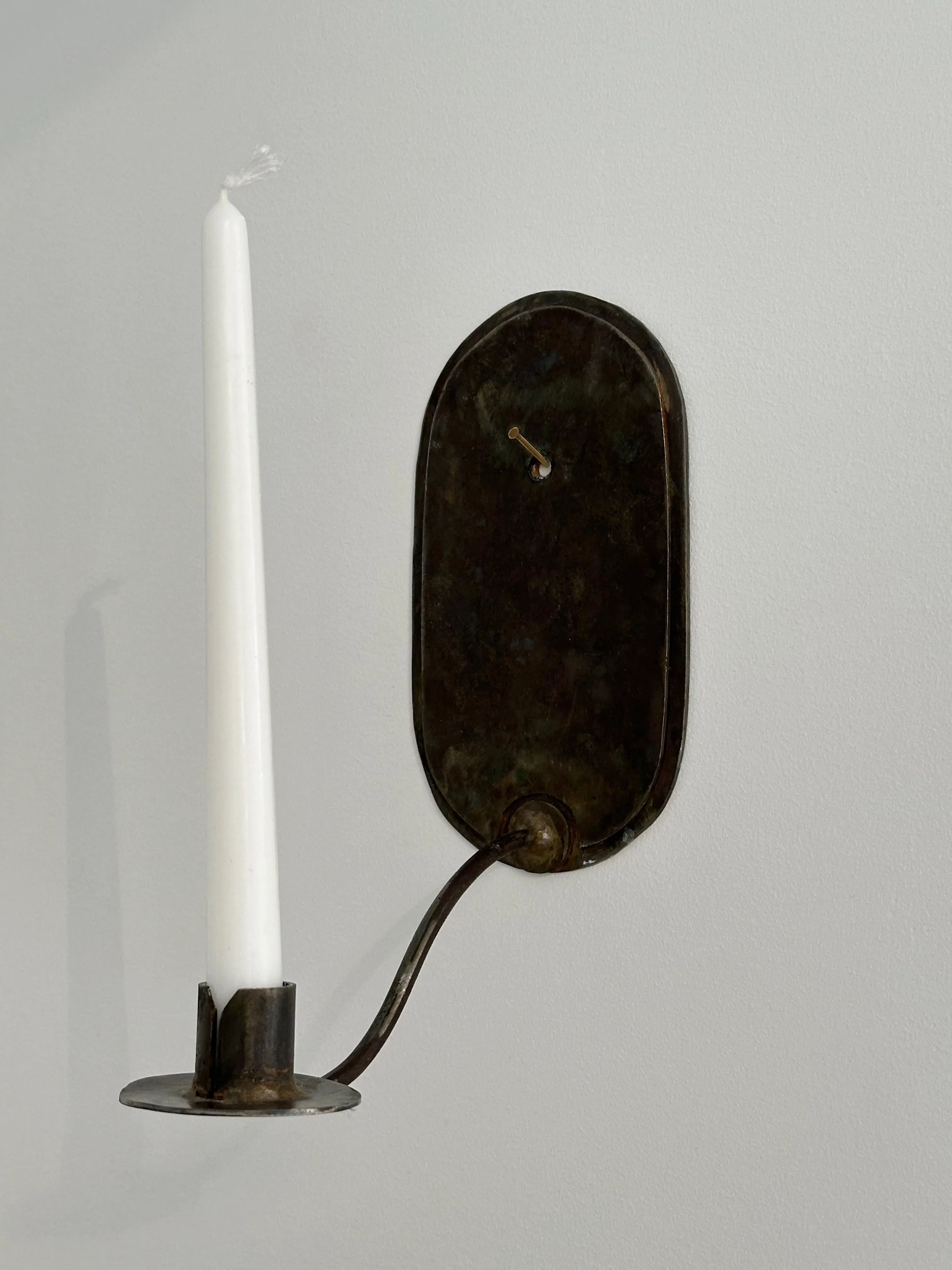 The Bronze Candle Sconce by Galerie Storm features a timeless silhouette with an oval backplate and elegantly curved arm, cradling a tall white candle. Its minimalistic design highlights masterful craftsmanship when mounted on a light-colored wall.