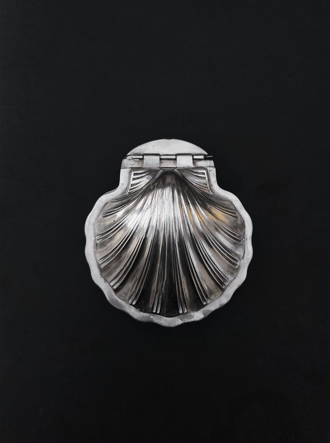 The Les Objoies Silver-plated Shell Box, scallop-shaped with a hinged lid, elegantly adorns a dark backdrop, embodying the charm of a decorative container perfect for sophisticated home decor.