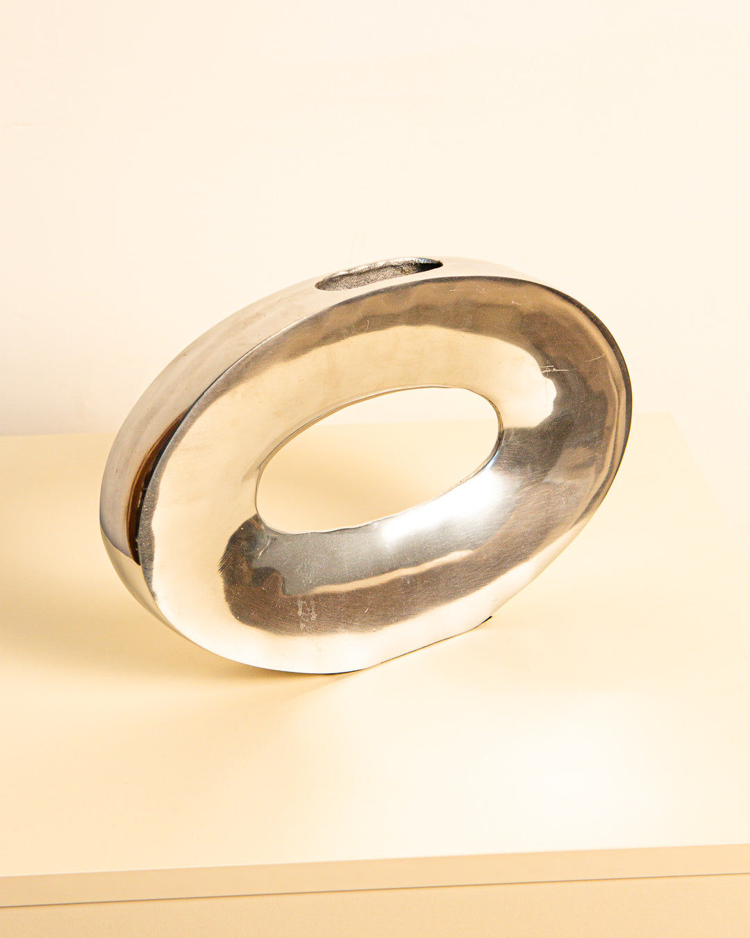 The "Ovale" Aluminum 80's sculpture by Treaptyque, a shiny oval piece with a hollow center reminiscent of vintage design, sits on a light surface against a plain background.