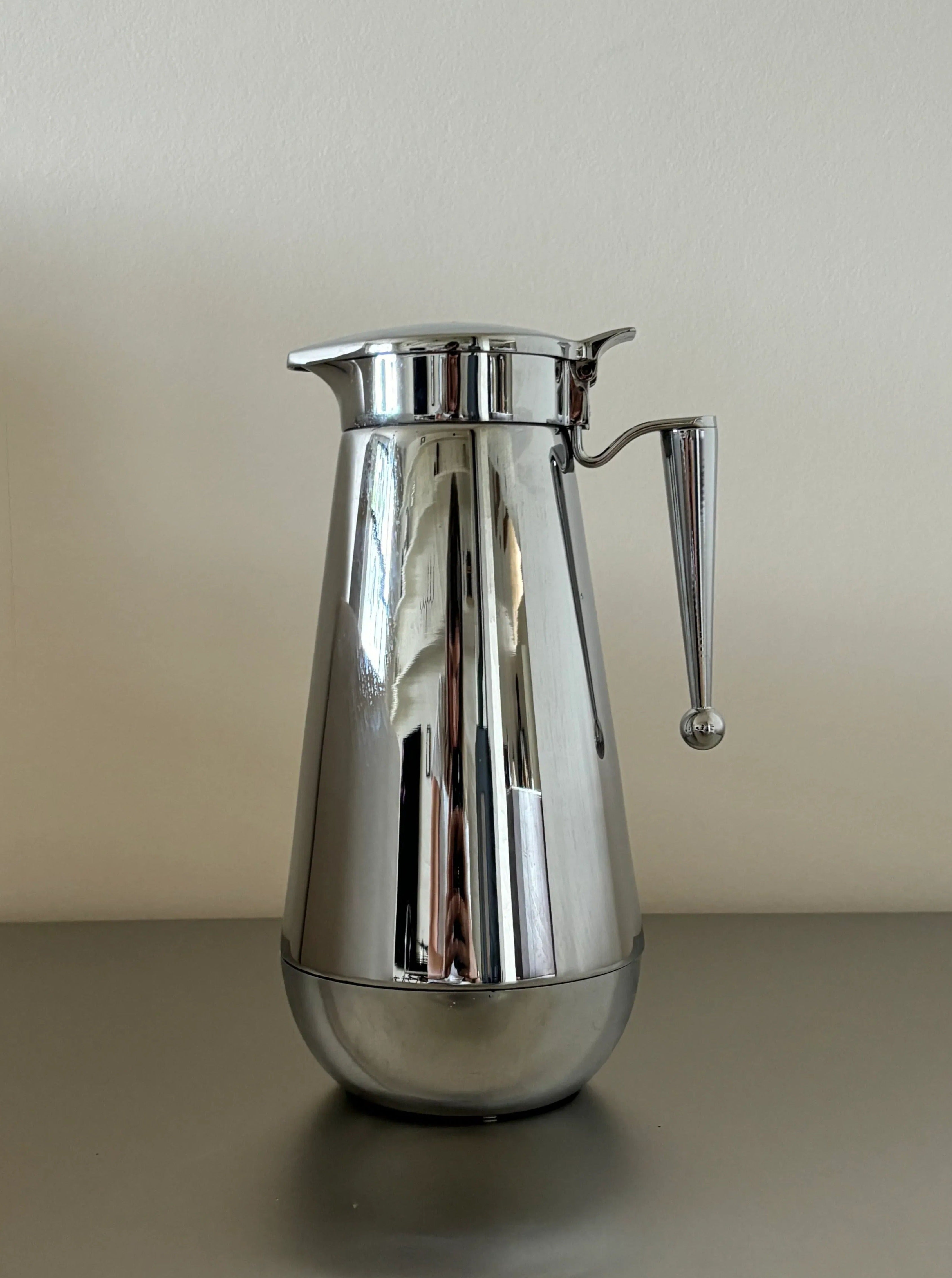 A sleek, stainless steel kettle in impeccable condition stands on a smooth, light gray countertop. Crafted by CU-RA-TED, the Steel Kettle "Erhard Germany" features a broad base that tapers towards the top, with a lid and handle adorned by a small spherical knob at the end.
