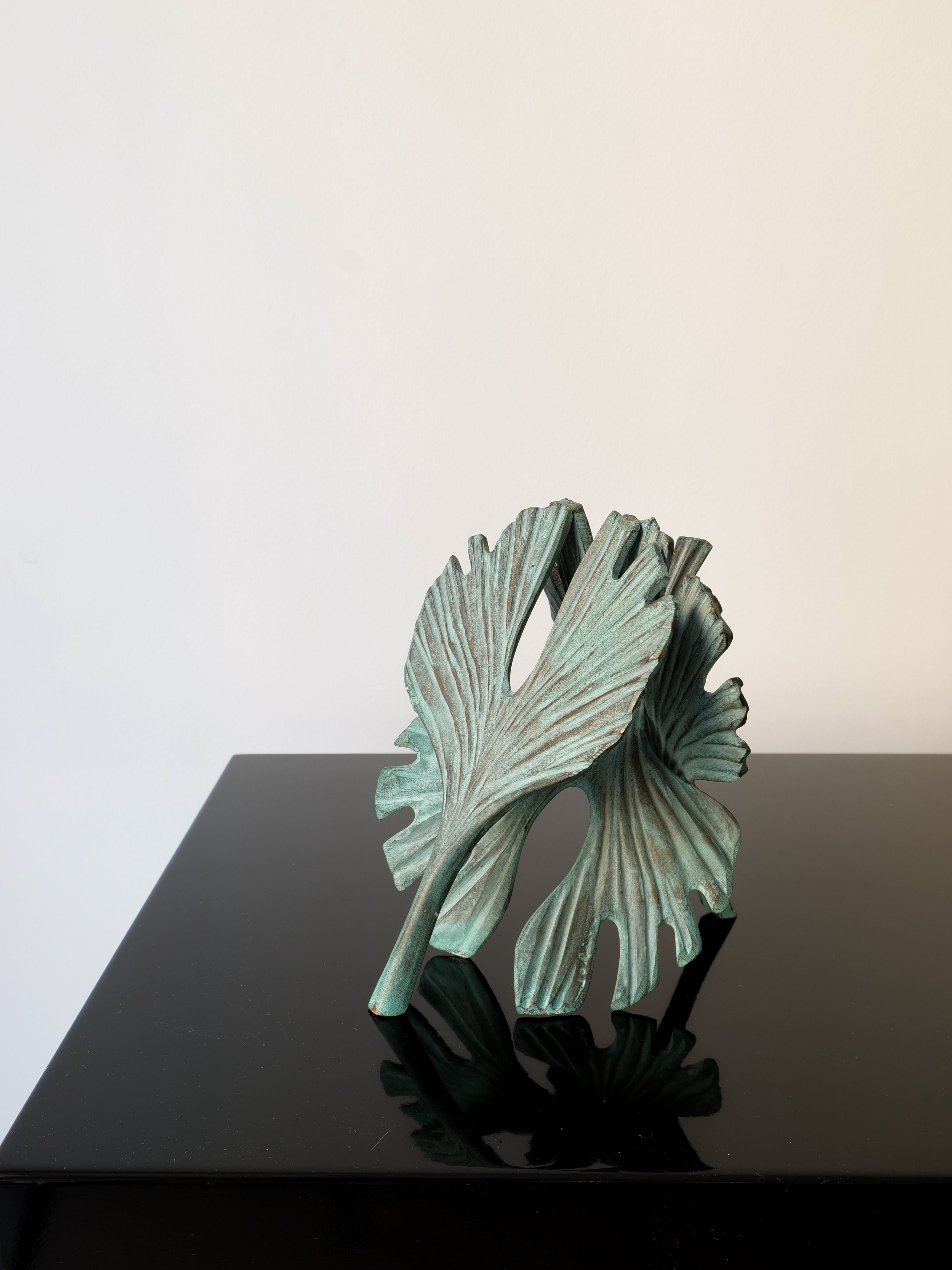 The Bronze Ginko Biloba Sculpture by Collection apart, with abstract intertwined leaves and a textured greenish patina, rests on a glossy black surface against a neutral backdrop.
