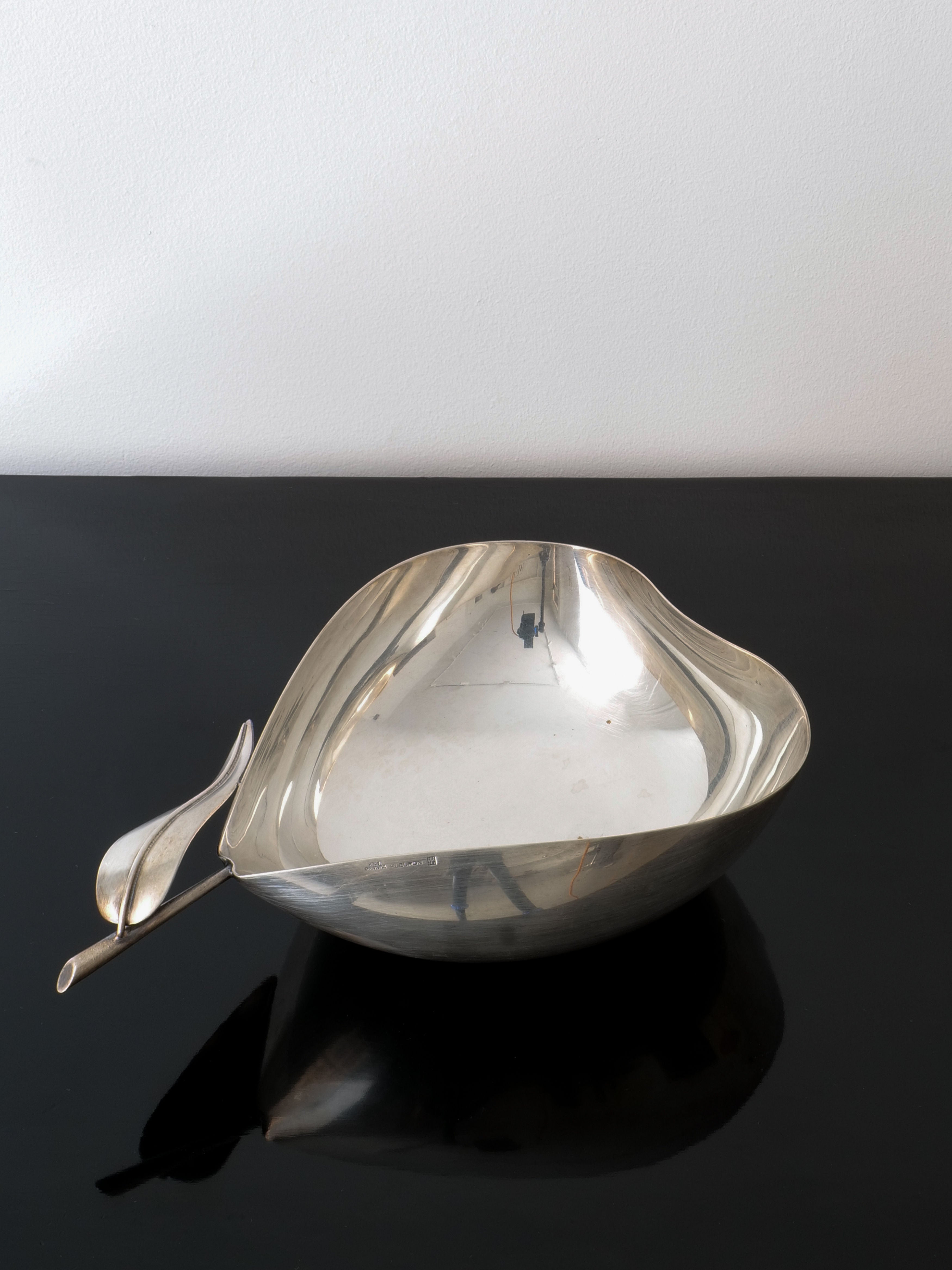The Christofle Vide Poche Lino Sabattini by Collection apart, known for its collaborations with Christofle, sits gracefully on a glossy black surface. Its asymmetrical design is highlighted by a distinctive wavy handle that resembles a leaf, set against the simplicity of a plain white wall.
