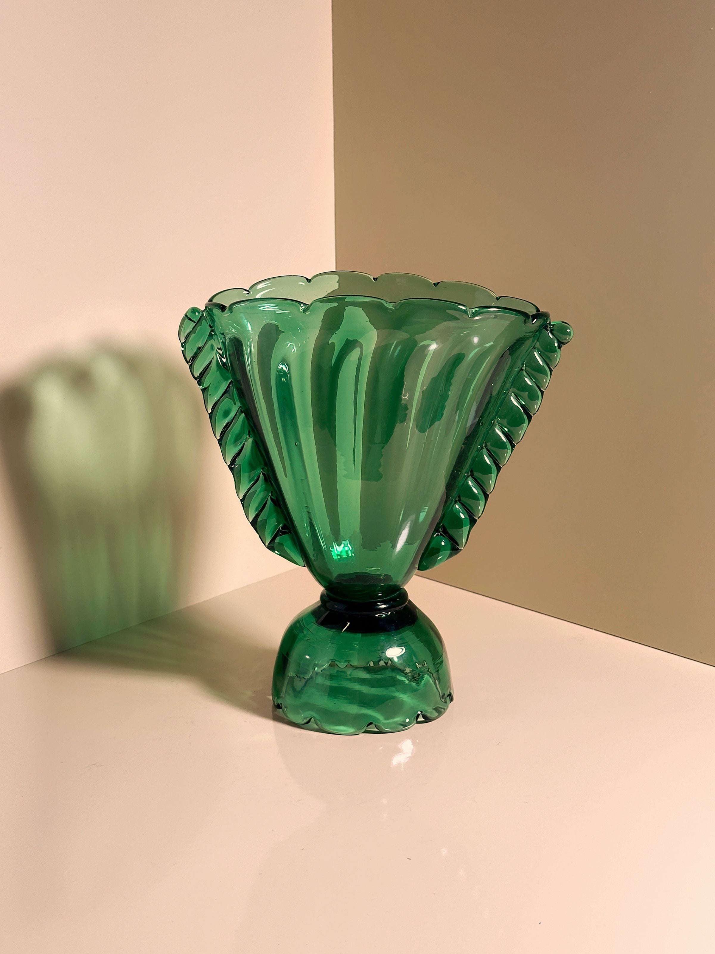 The Glass Amphora Vase -1910s by Dodo Vintage features a scalloped rim and leaf-like handles. This handmade green glass vase casts a soft shadow on a plain, light background, highlighting its translucent and glossy look.