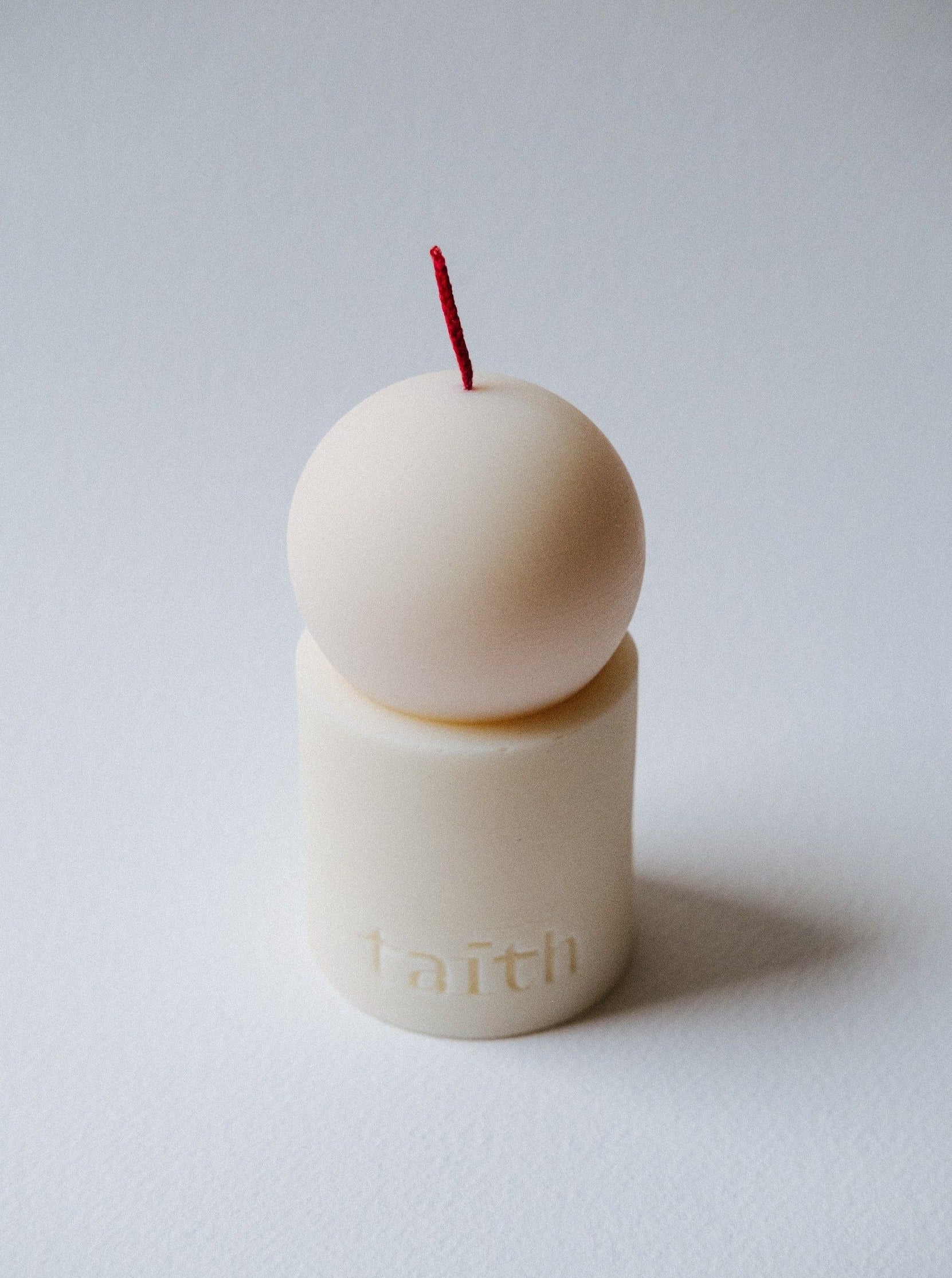 A minimalist off-white Taith Lulu Candle Cream with a red wick is positioned on a white surface. The sculpted soy candle features a spherical top and a cylindrical base, with the word "faith" embossed on the base. Its simple and elegant design pairs beautifully with its captivating fragrance.