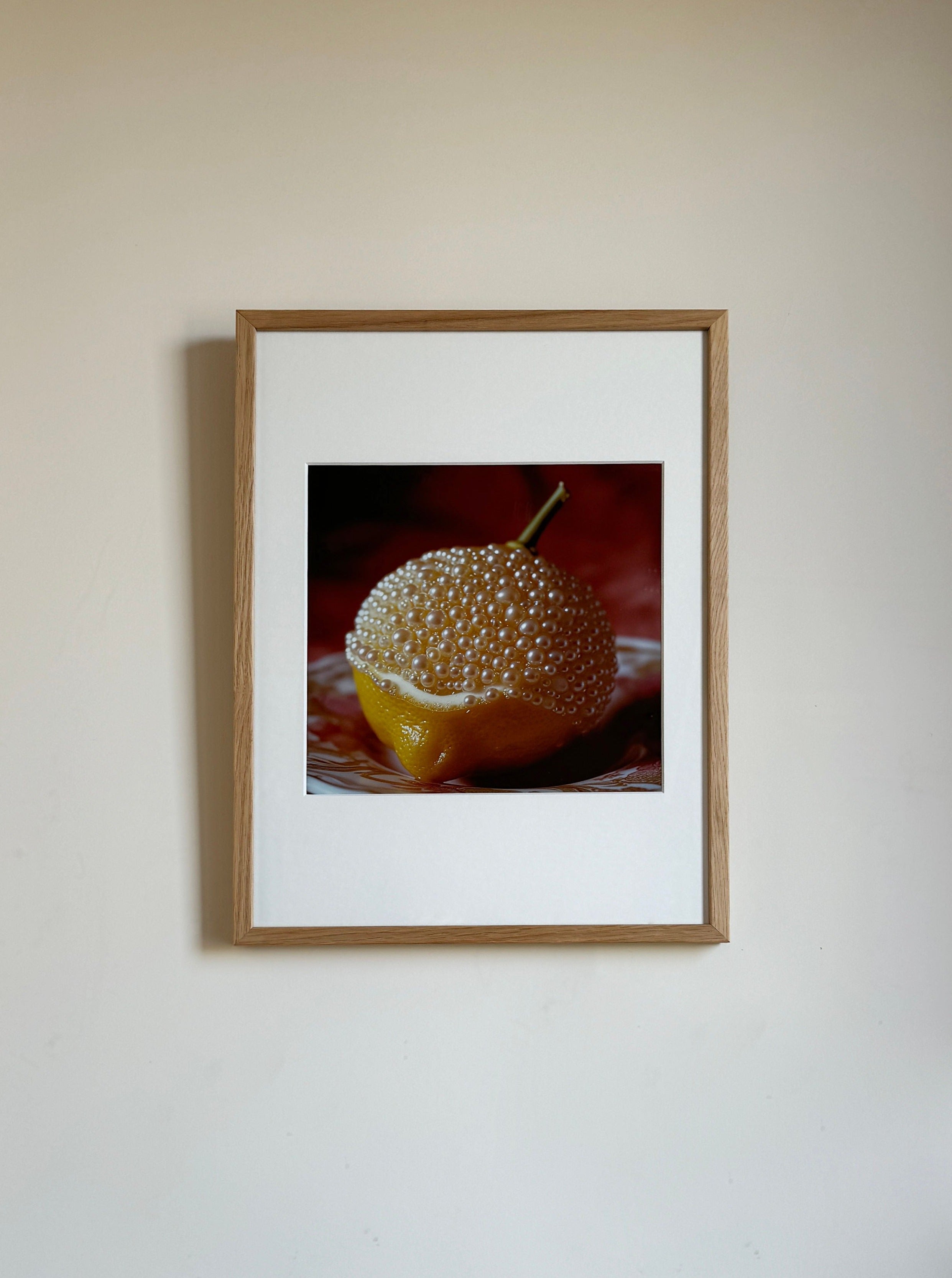A framed photograph hanging on a plain wall, depicting a close-up of a half lemon covered in clear, bubble-like spheres. The Lemon and Pearls by Adeline Mai combination is set against a dark, textured background. The wooden frame surrounds a white mat with the mixed media photography centered perfectly.