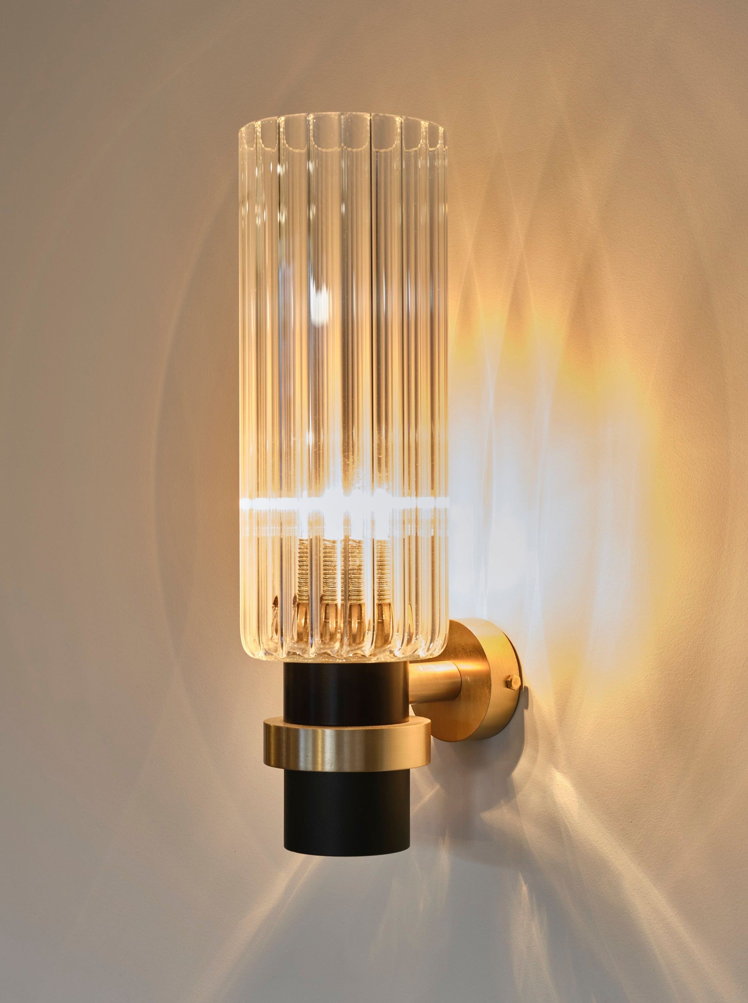 A modern dimmable Marine Breynaert Magda I Wall Light with a cylindrical, clear glass shade and golden accents illuminates a warm yellow light, casting soft shadows on a light-colored wall.