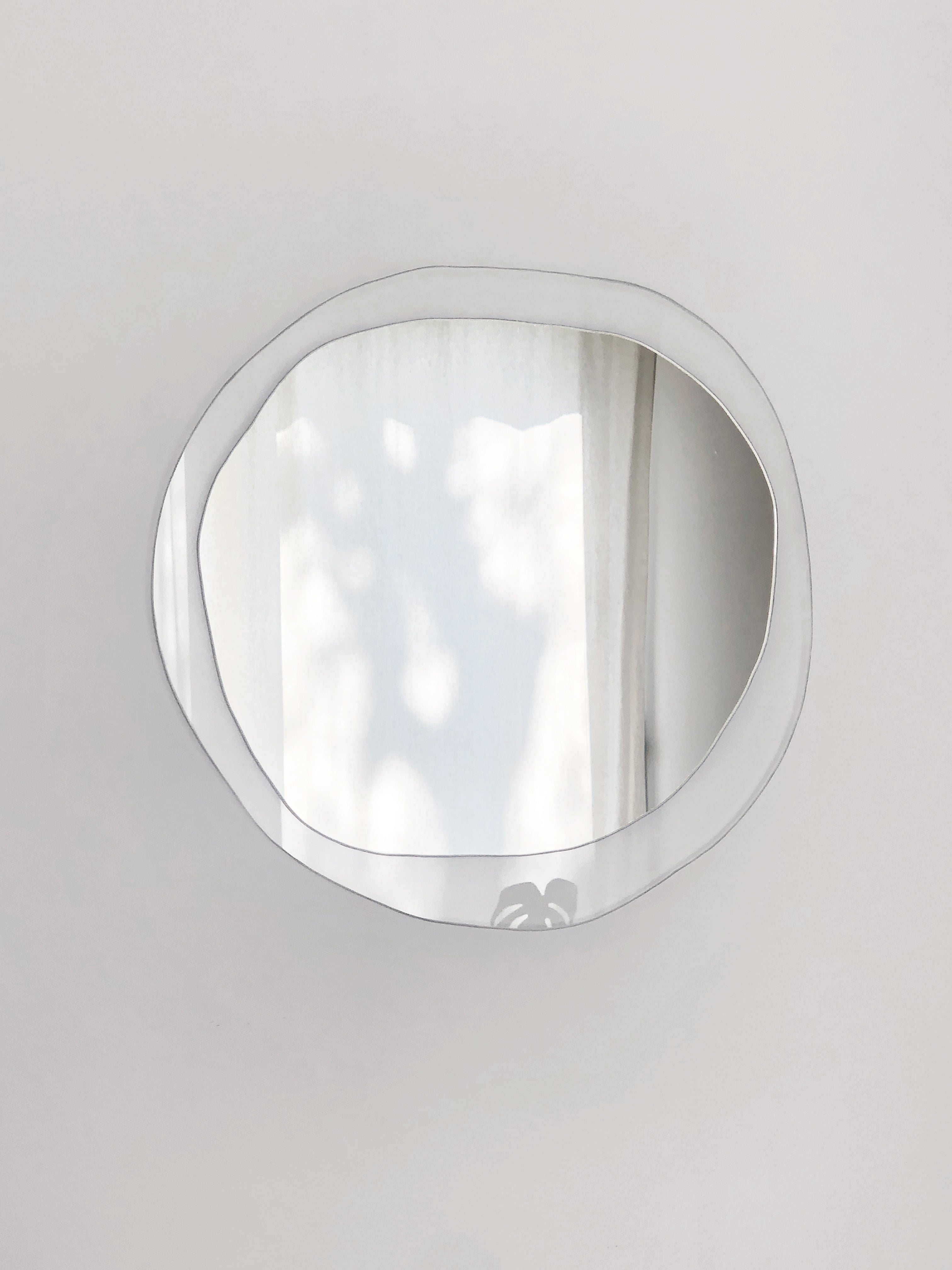 The irregularly shaped Mirror Ombrée Clair by Laurène Guarneri beautifully reflects light, casting shadows of leaves onto a light-colored curtain in the background. Its organic shape introduces a modern, artistic flair to the minimalistic setting, highlighting its unique composition.