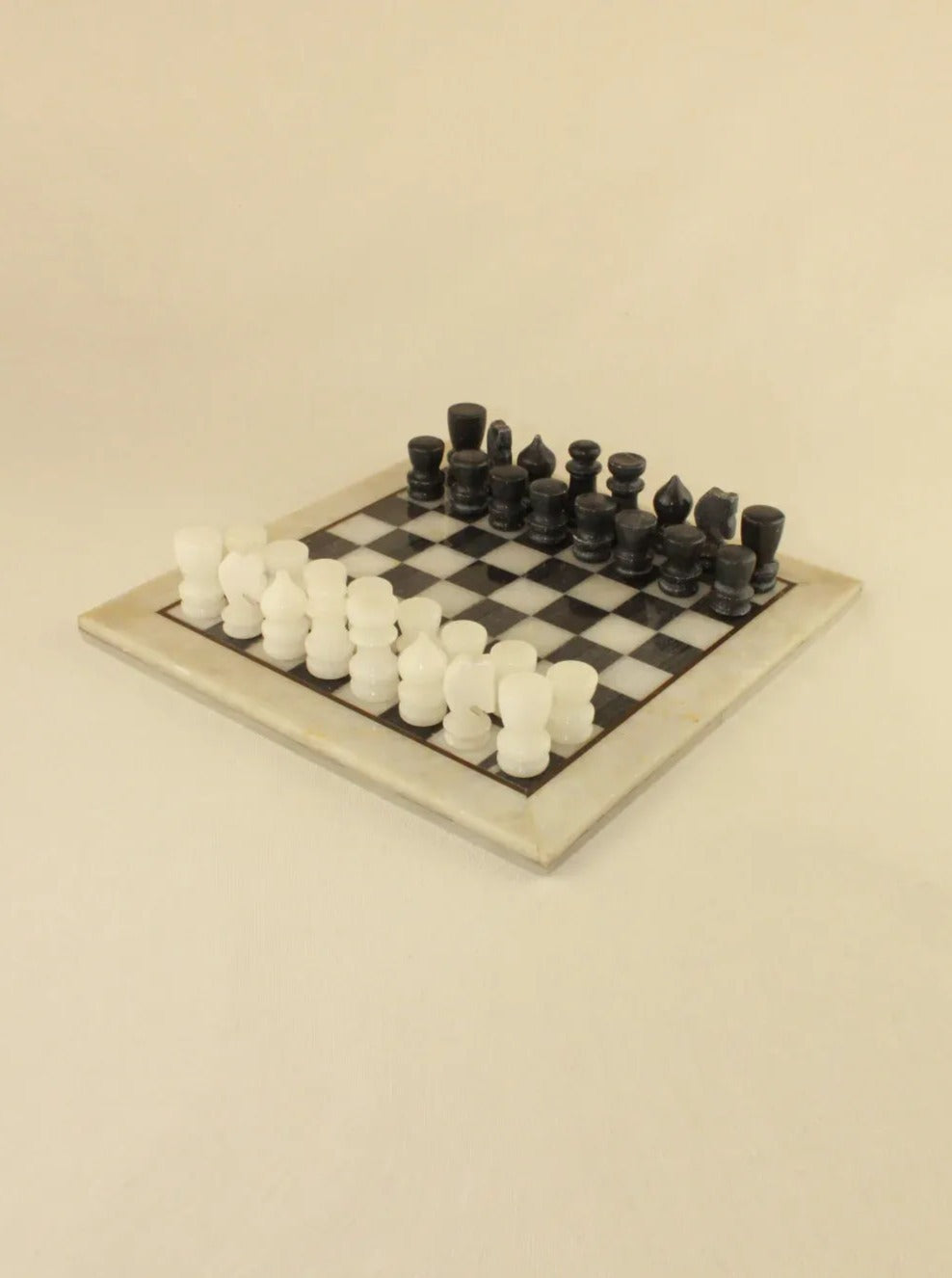A Marble Chess Board by Boga Avante Shop is set up for a game, with all pieces in their starting positions on a beige background. The board has alternating light and dark squares, and the hand-sculpted pieces are