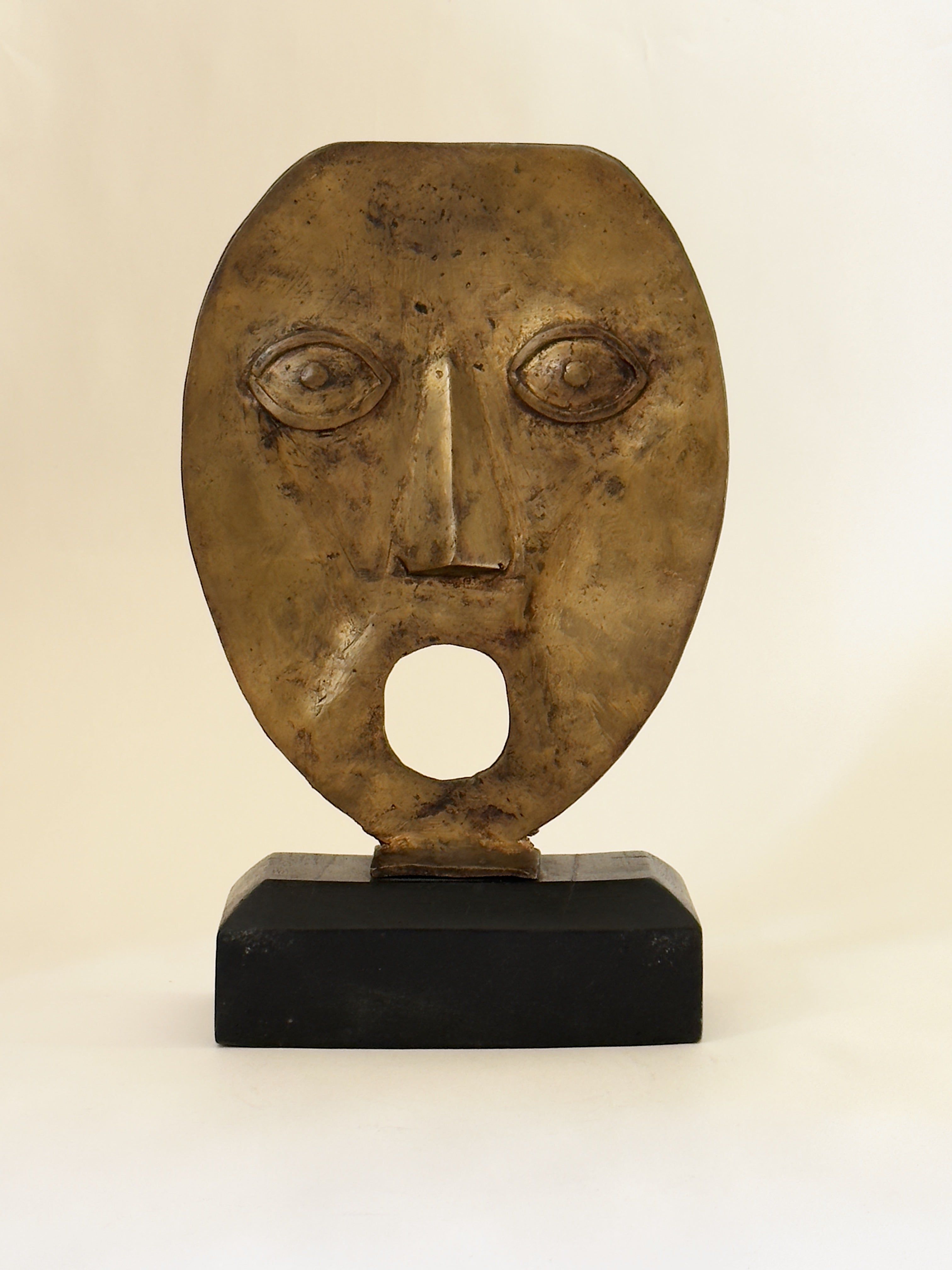 The Galerie Storm Brutalist Bronze Face on Wooden Base features wide eyes, a prominent nose, and an open mouth, displayed on a black rectangular base against a plain light background.