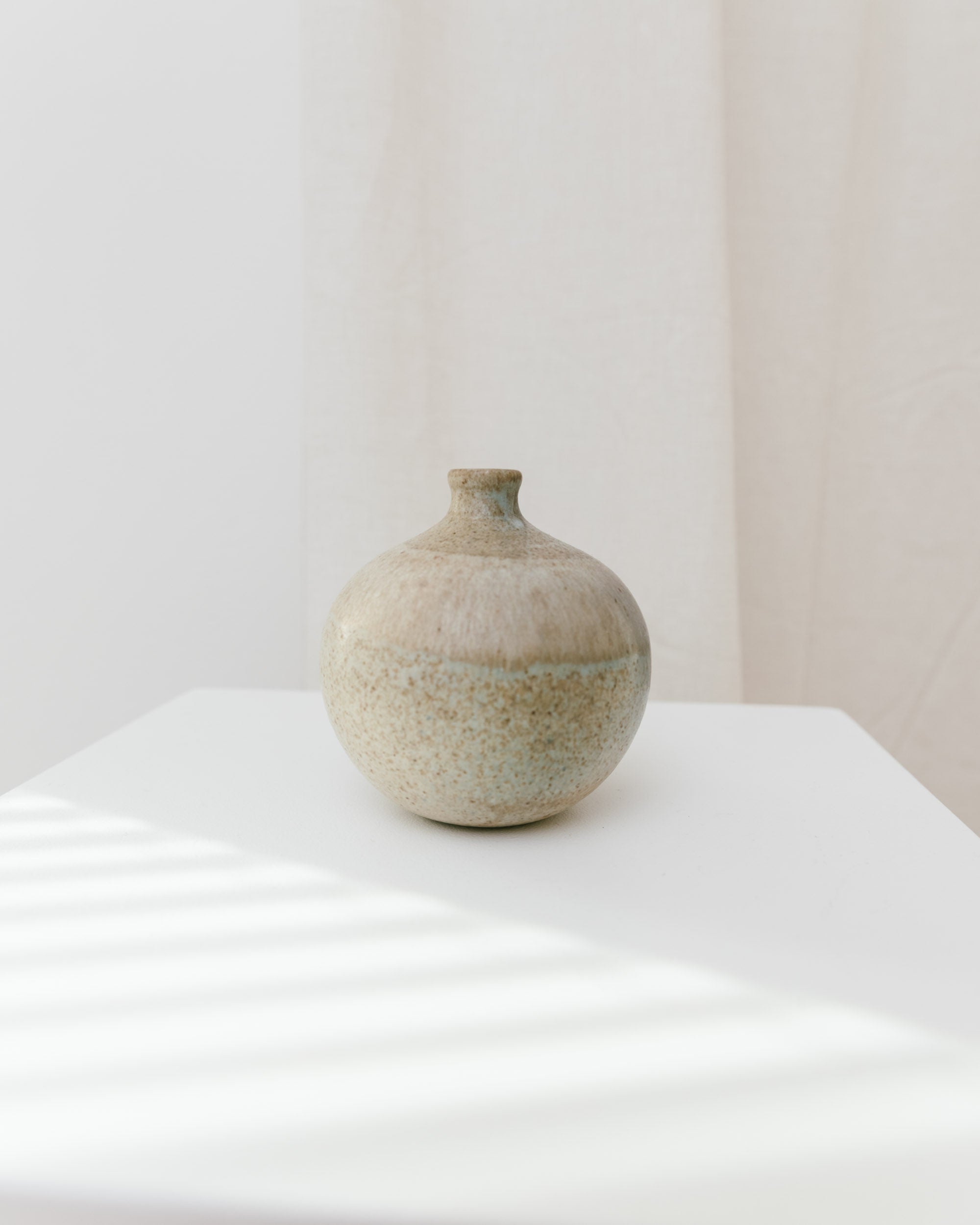 A small, round beige Ceramic Vase by Bernd Röter 1970s from Bottega Jacobs sits on a white surface. Soft sunlight casts diagonal shadows, while a light neutral curtain background adds minimalistic charm with a hint of mid-century design elegance.