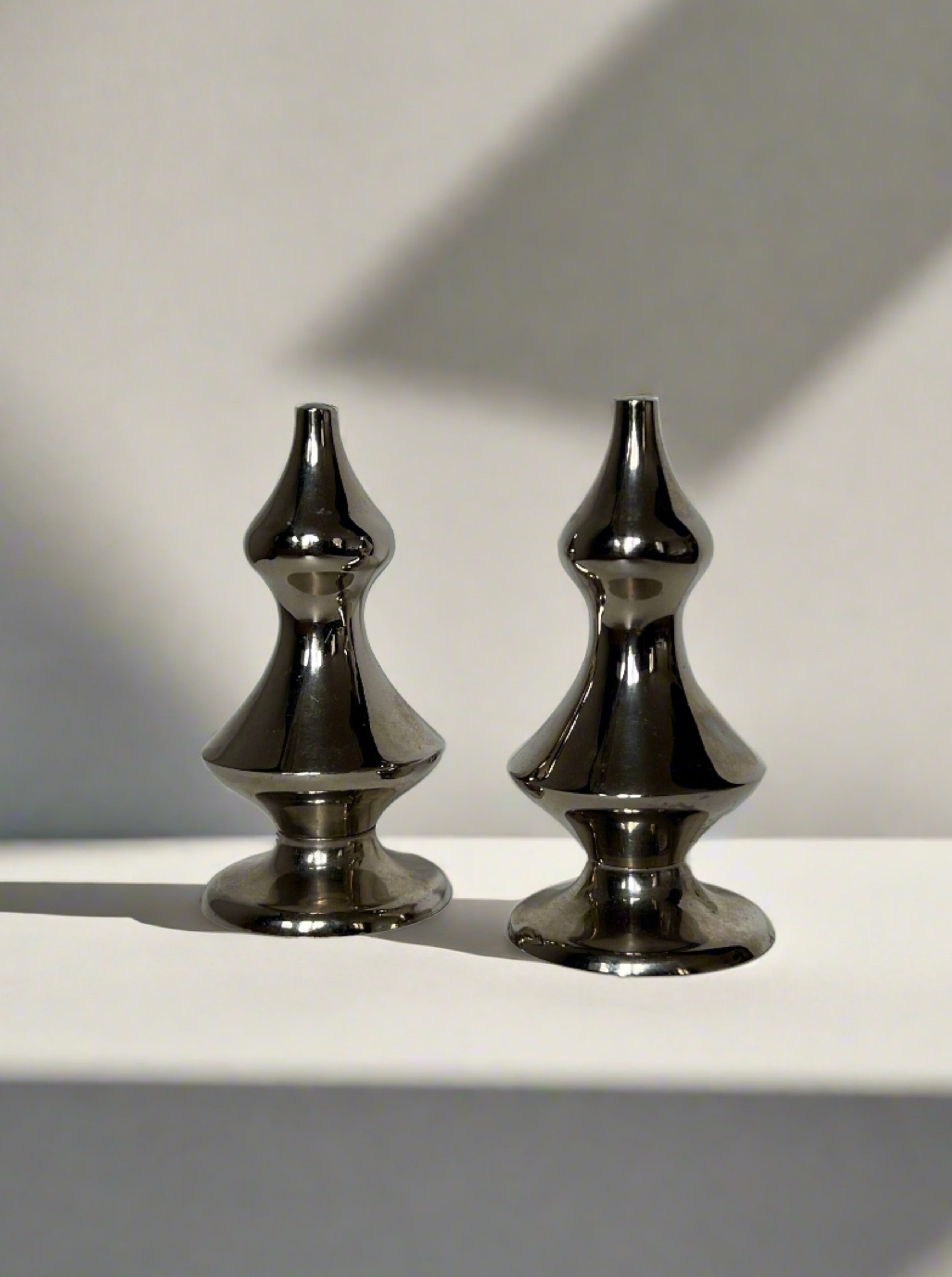 A pair of silver-plated, elegantly shaped Art Deco Salt & Pepper shakers with a contemporary, geometrical design by Manushi, standing side by side on a white surface against a plain background.