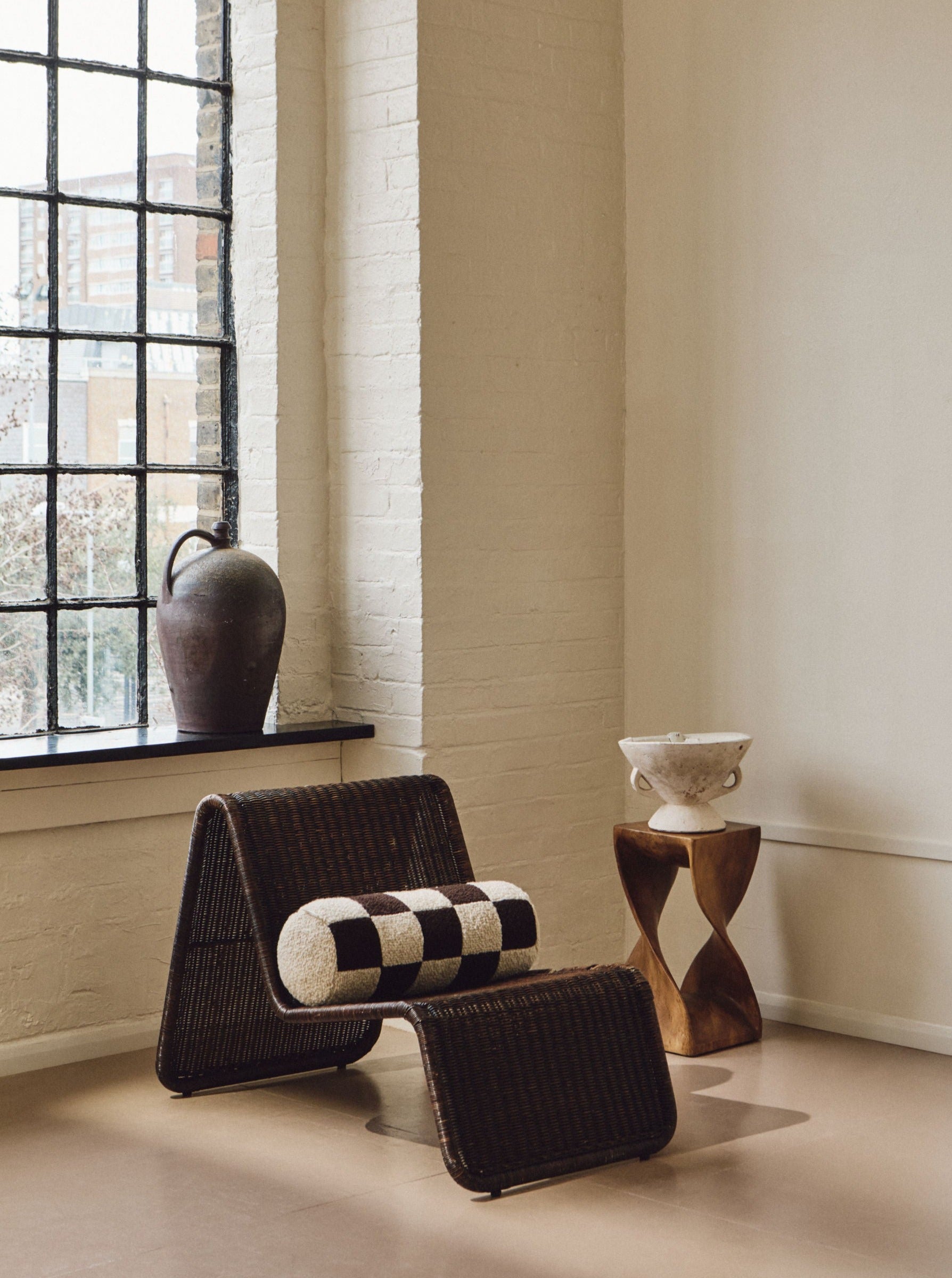 A cozy corner features a wicker chair with an ottoman, adorned with a vintage-inspired cushion made of British eco wool. A wooden accent table beside the chair holds a decorative bowl. A large, rustic pottery vase sits on the windowsill of a tall, industrial-style window, while the cushion is replaced by AMÉLIORÉ's Le Cylindre - Wool Bouclé - Chocolate/Eggshell for an added touch of elegance.