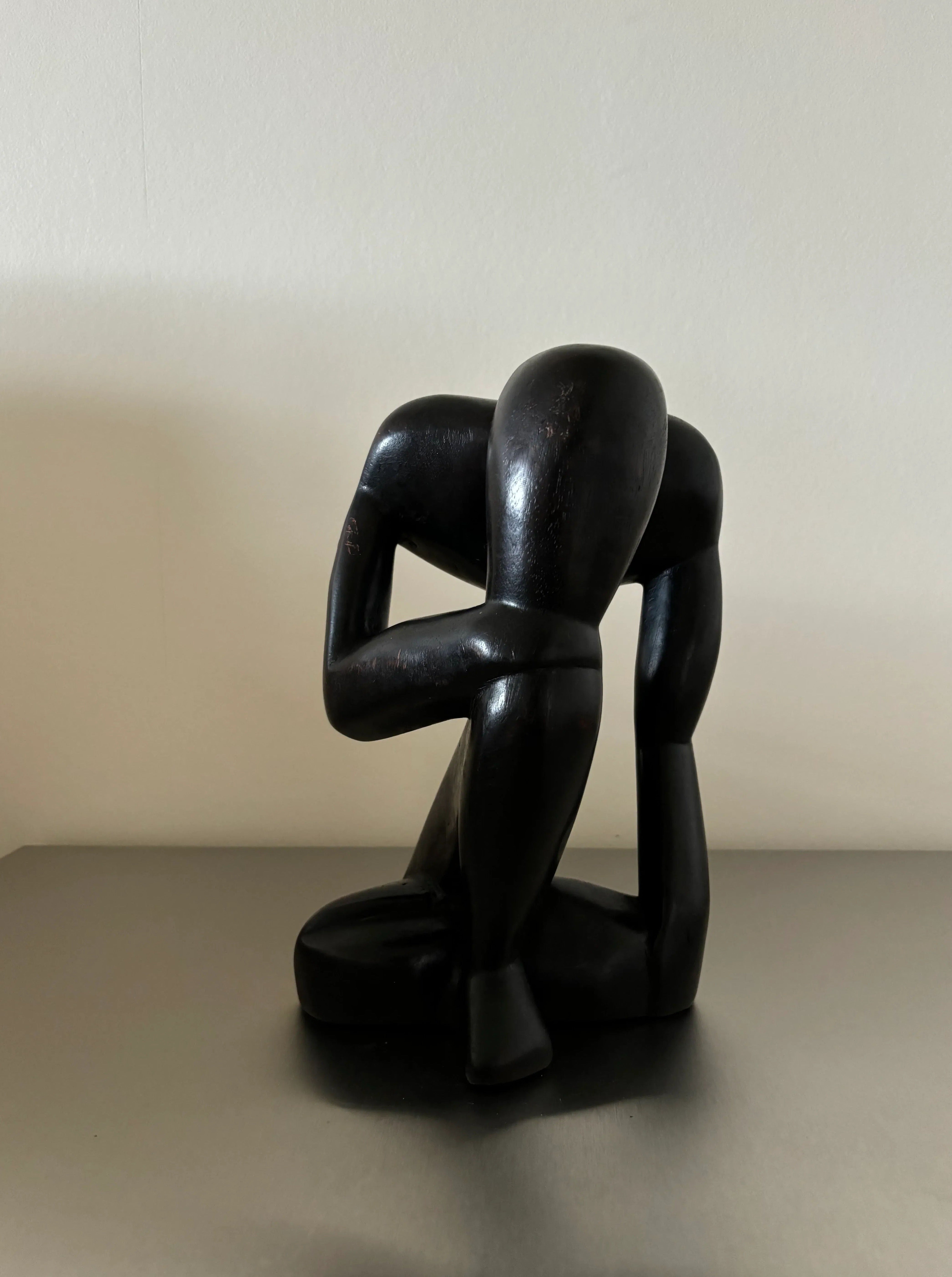 A dark, abstract wood sculpture of a figure kneeling down with its head resting on its arms. The piece showcases fine craftsmanship with minimalistic and smooth lines, complemented by a black finish, and is placed against a plain, light-colored background. Introducing Black Wooden Object - “The Thinker” by CU-RA-TED.