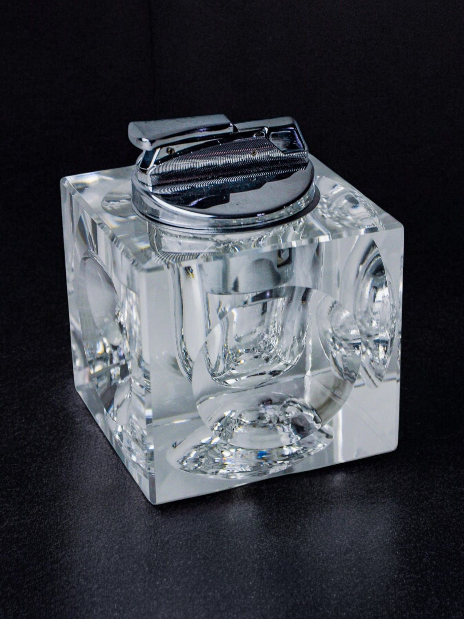Displayed against a black background is the Ice Cube Lighter by Antonio Imperatore 70s from Malamar Studio, featuring a transparent, cube-shaped design with a metallic top. This exquisite piece creates intricate reflections and patterns reminiscent of Murano glass crafted by Venetian master glassmakers.