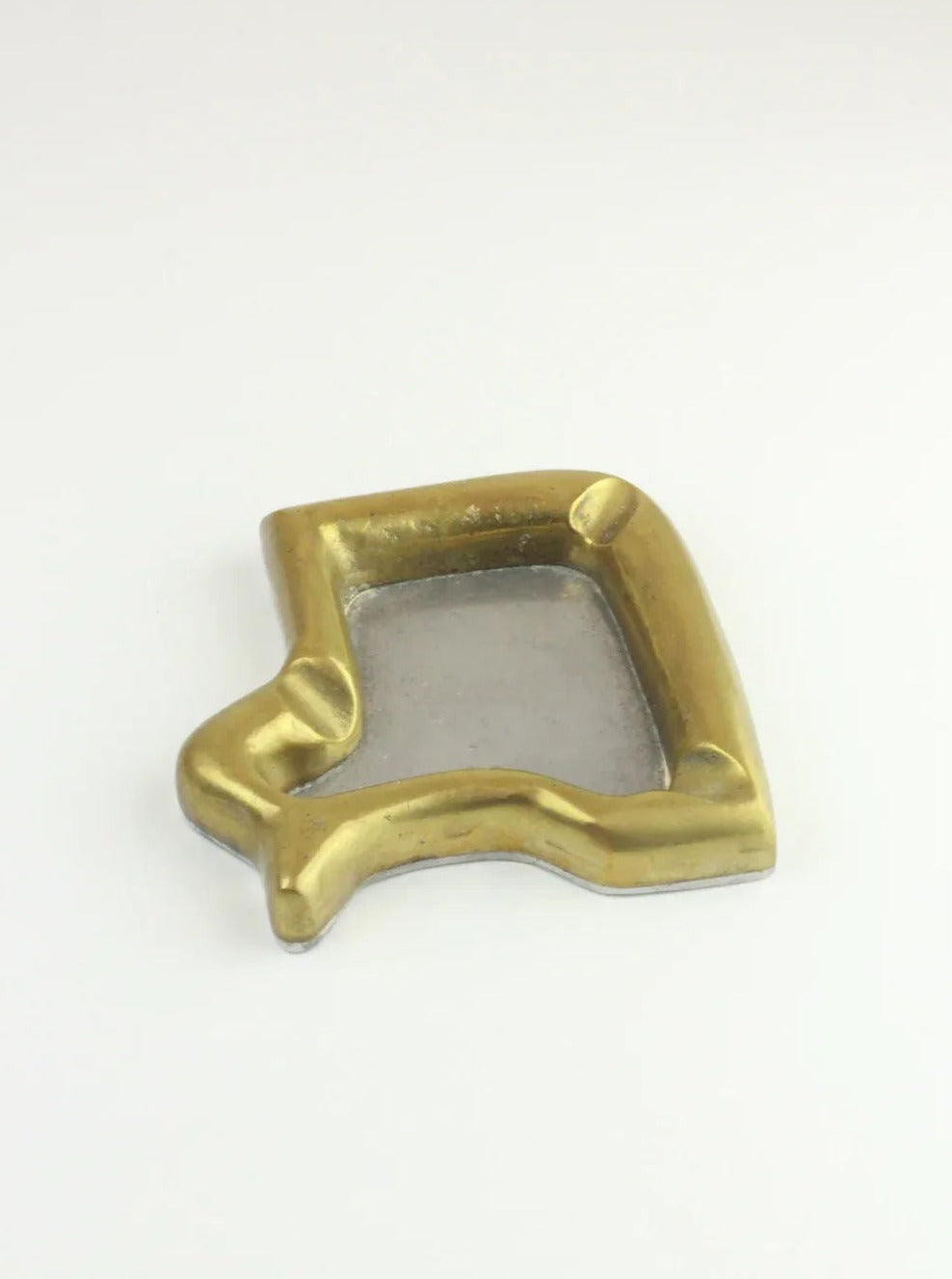 An abstract-shaped metal dish with a slightly raised perimeter and an empty, smooth center evokes the essence of a Boga Avante Shop piece. This gold-colored dish, composed of brass and aluminum, named the Brutalist Ashtray, features a modern, sculptural design reminiscent of Brutalist ashtrays and rests elegantly on a white surface.