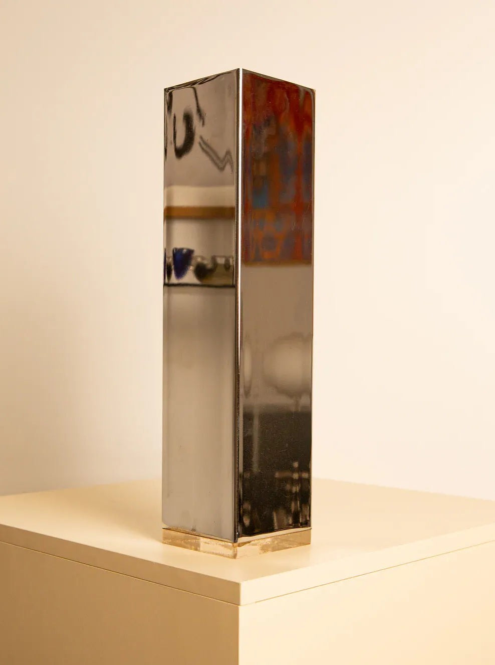 A tall, reflective rectangular prism sculpture, reminiscent of a Large "Colonne" Vase 70's by Treaptyque, stands on a beige pedestal against a plain background. The mirrored metal surface showcases its surroundings with a blend of metallic and glossy textures, evoking an elegant 70s design.
