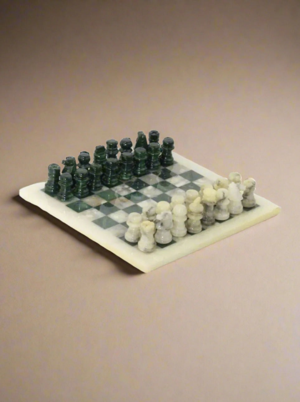 A *Boga Avante Shop* *Vintage Green and White Marble Chessboard* with elegant green and white pieces is set up for a game. The green pieces are on the left, and the white pieces are on the right, placed on a vintage chessboard oriented so that the white square is in the bottom right corner. The background features a plain surface.