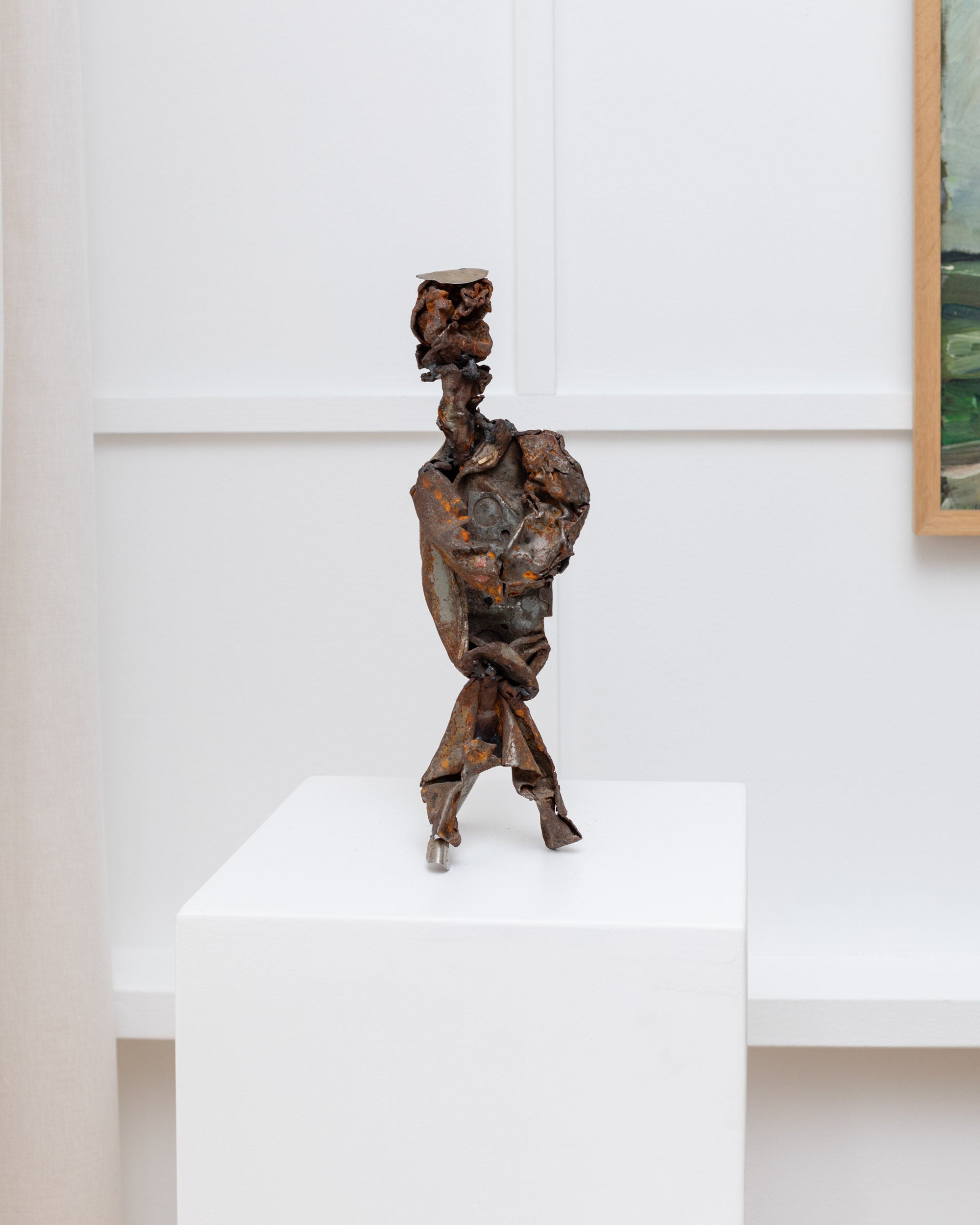 A small, handmade metal sculpture by Bottega Jacobs stands on a white pedestal in a minimalistic gallery. This vintage piece from 1970s Italy features twisted, rusted metal forming an abstract humanoid figure with head, torso, and legs against a white wall with framed art partially visible.
