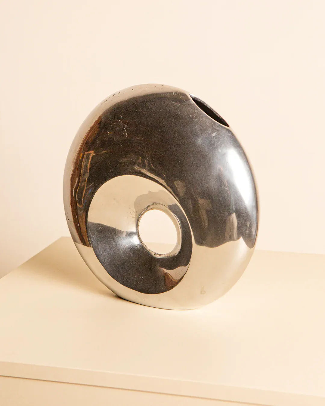 A reflective abstract circular sculpture, similar to the "Cercle" Vase in Aluminum 80's by Treaptyque, features a central hole. Its metallic finish distorts reflections on a beige surface, while the simple background accentuates its unique shape and smooth texture from a private collection.