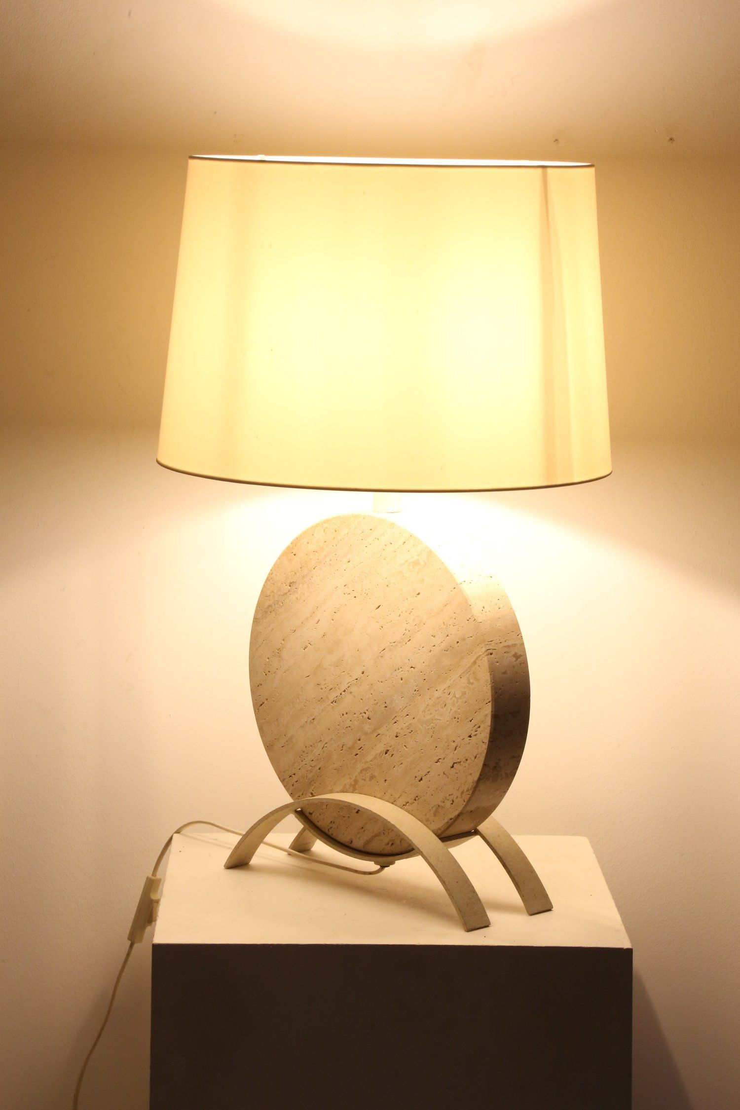 The Ancien et Jolie Italian Travertine Table Lamp boasts a sculptural design with a beige finish, featuring a large, circular stone base and arched supports on a white stand. Its light cream lampshade casts a warm glow, accentuating the lamp's clean lines and elegance.