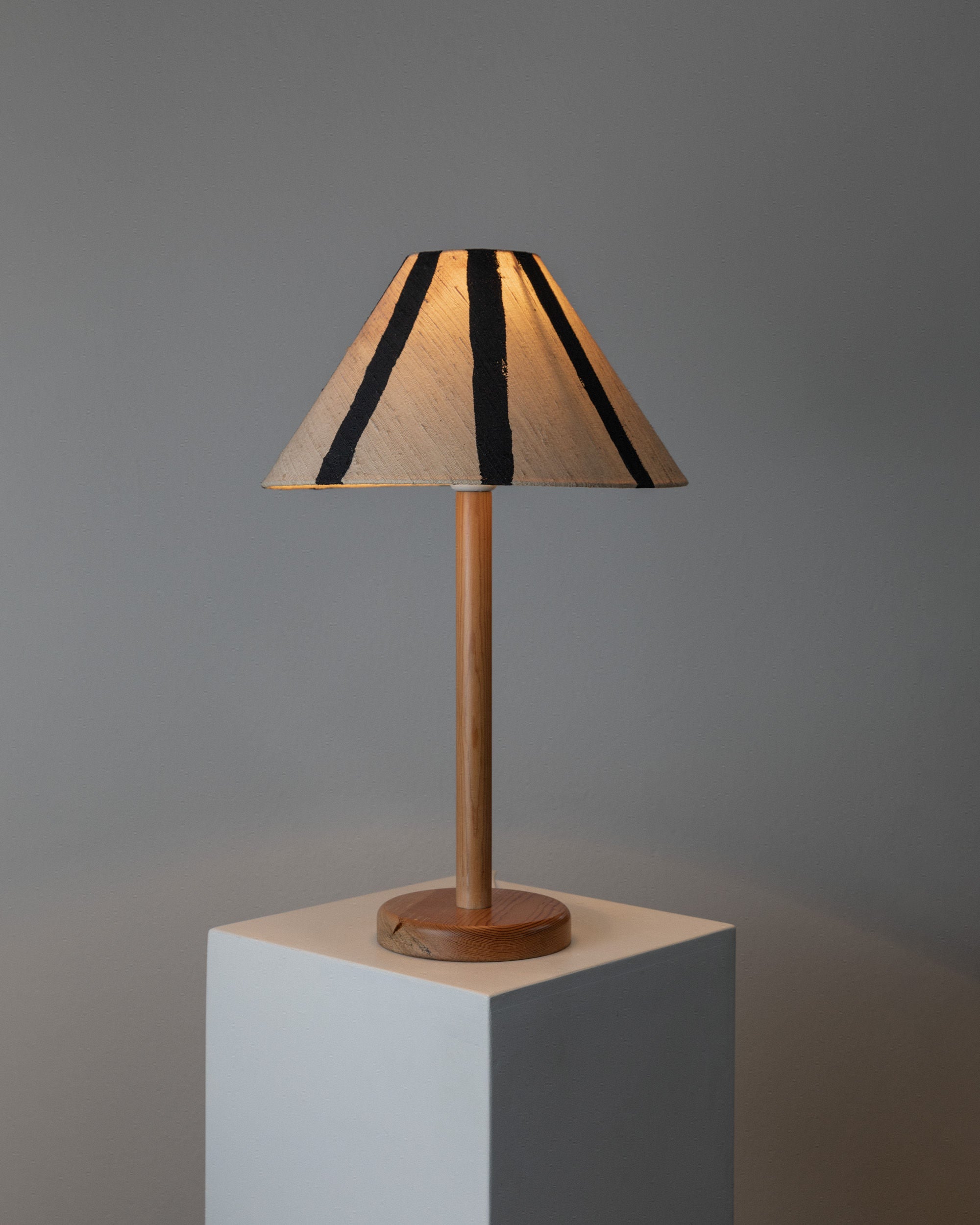 The Table Lamp by Solbackens Svarveri from Bottega Jacobs features a vintage design with a wooden base and hand-painted linen shade of black stripes on beige. Set on a white pedestal against a gray wall, it emits warm light echoing Swedish mid-century style.