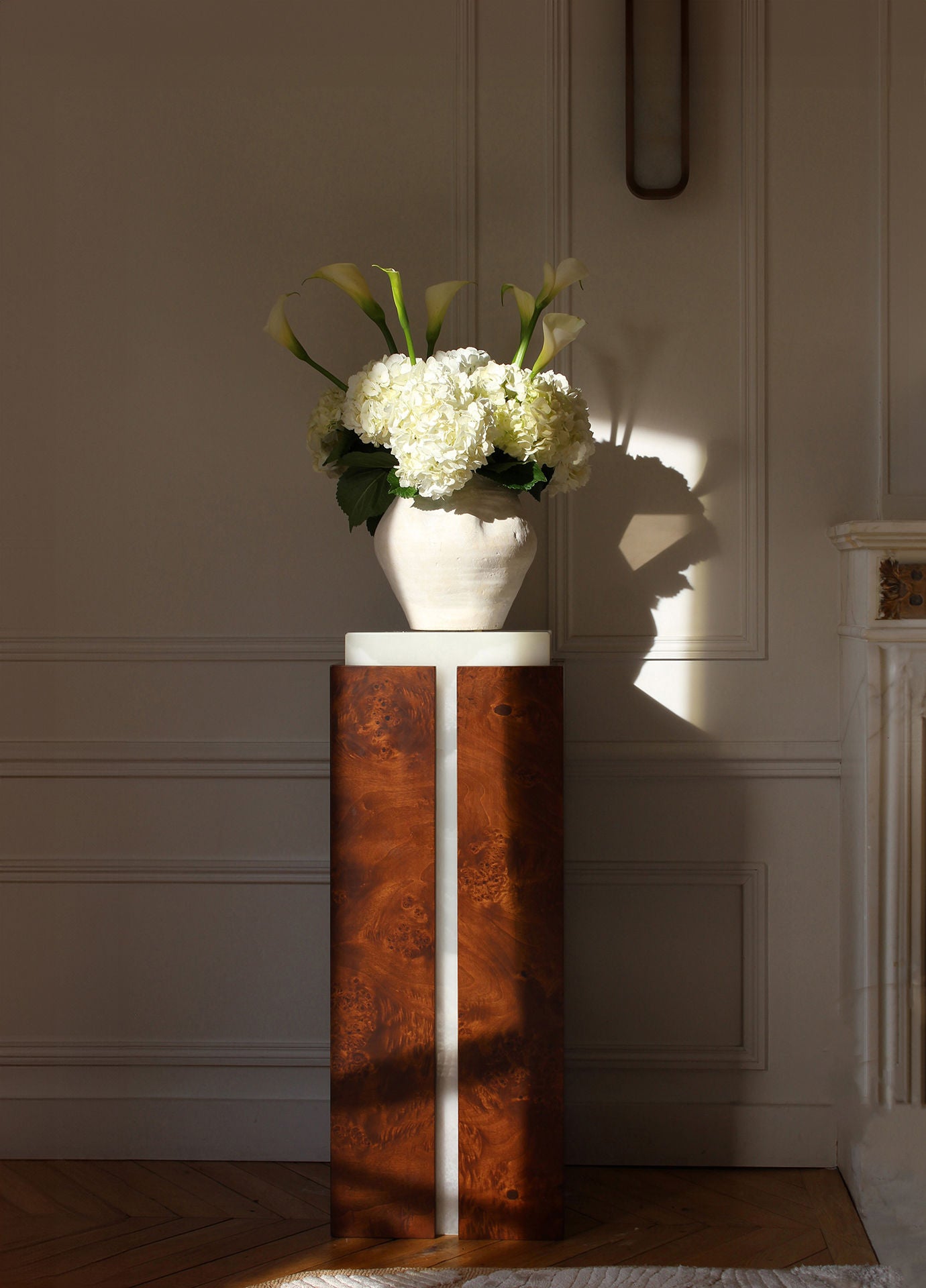 A sunlit room features the Marbera 'Como' Plinth in white onyx and brown wood, topped with a ceramic vase of white flowers. Soft shadows fall on a white wall with molding, while a brown wood fireplace peeks in from the right.