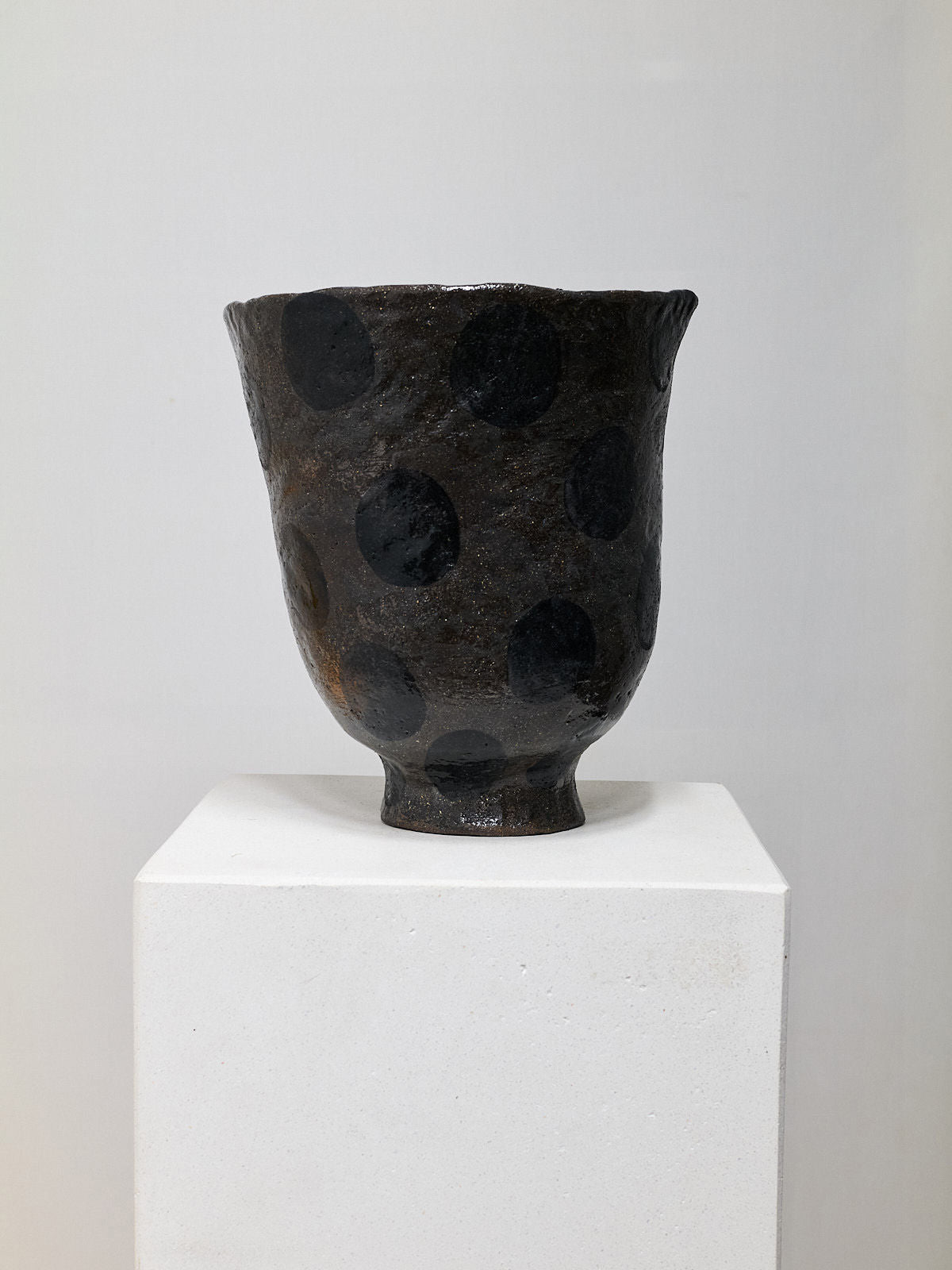 A Stoneware Vase by Claire Pain, featuring a dark textured design with a wide brim and bold black polka dots, is showcased on a white pedestal against a plain backdrop.