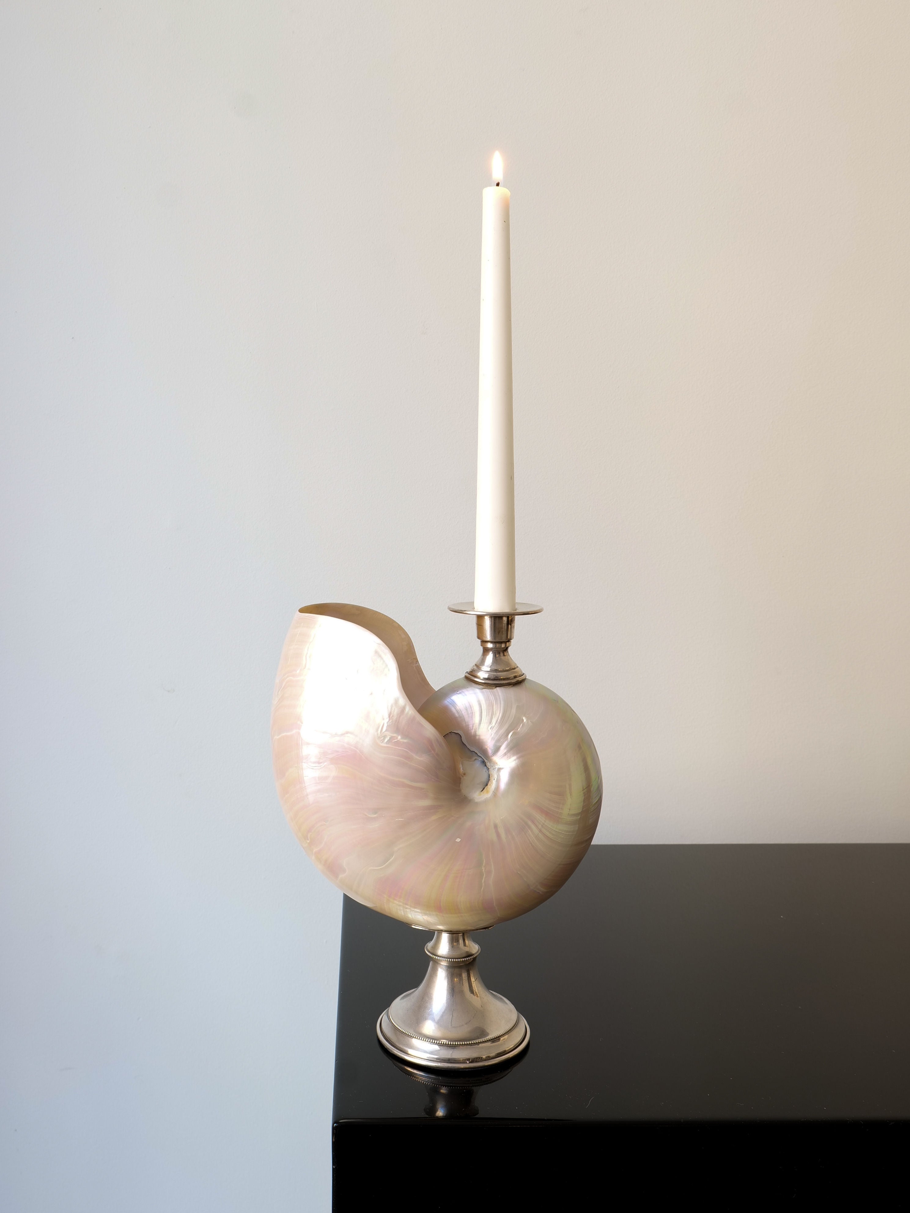 A white candle flickers in the Nautilus Silver Candle Holder by Collection apart, beautifully crafted from a nautilus shell. It rests elegantly on a shiny black surface against a plain light-colored background, exuding sophistication.