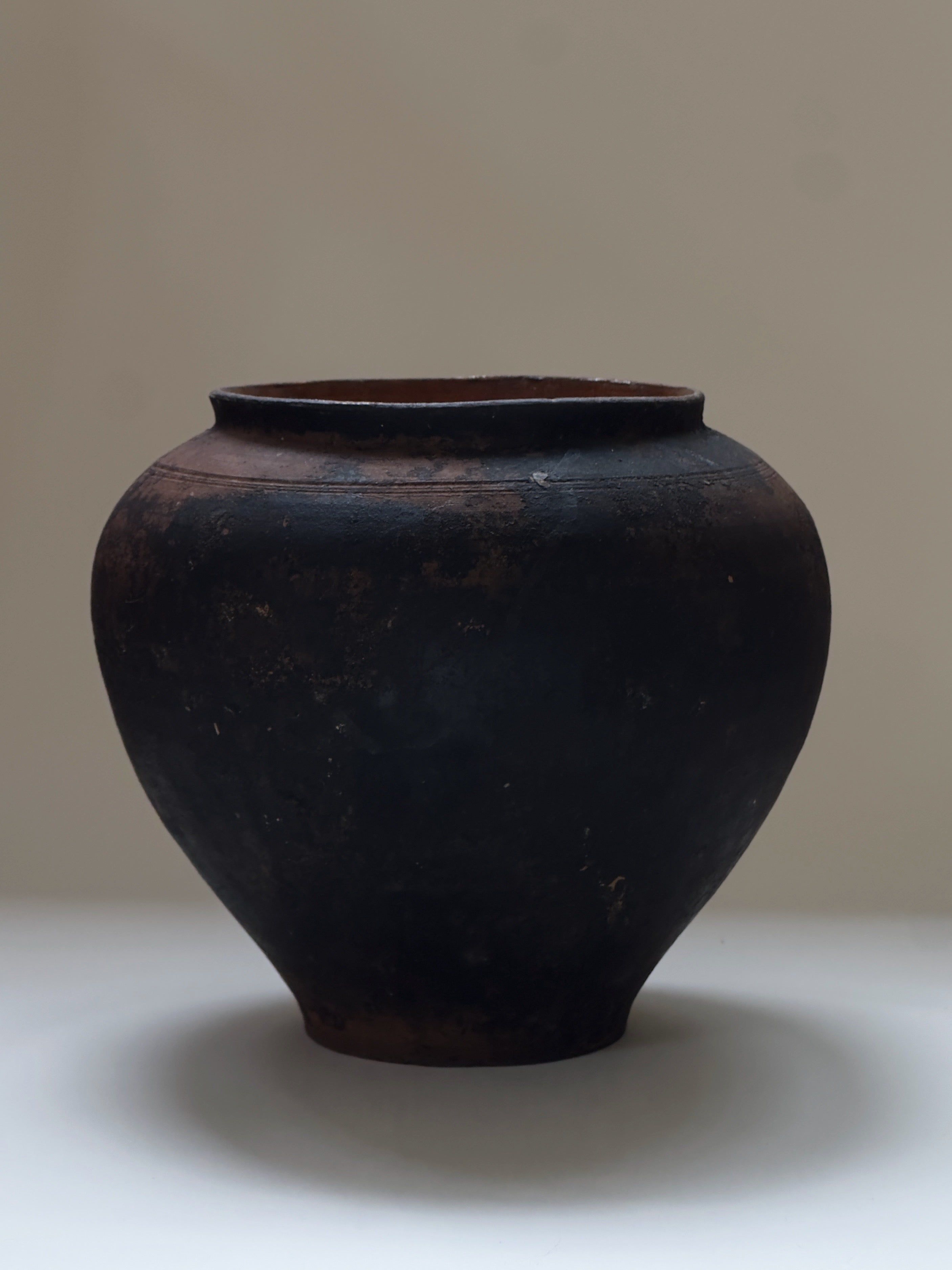 The Antique Vase by Galerie Storm is a large, round, dark piece with a narrow opening and rustic, textured surface, featuring a beautiful aged patina. It sits on a white surface against a plain, light background.
