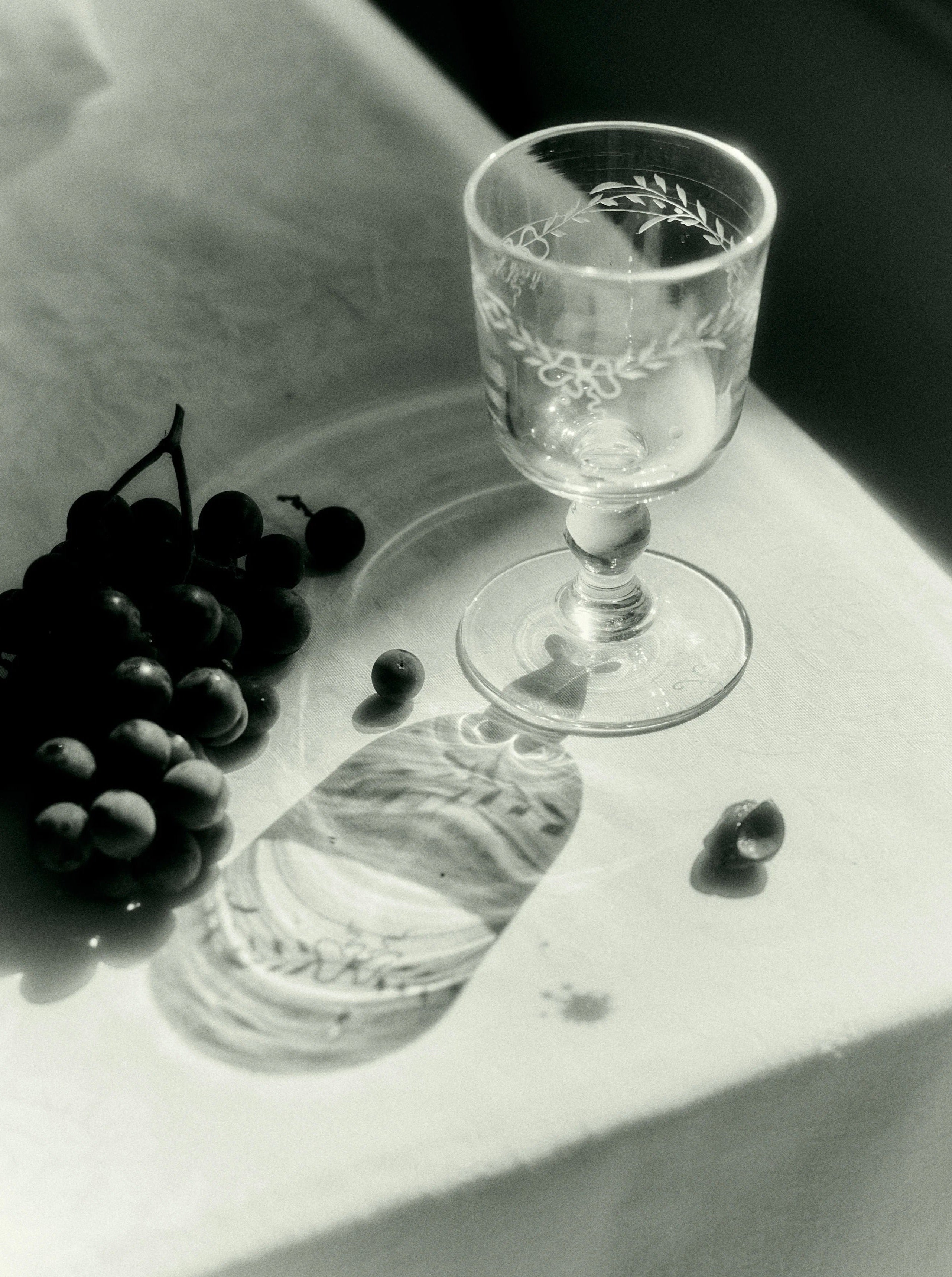 Black and white photo of a cluster of grapes next to an ornate Akua Objects Barbro Wine Glass - Set of four on a table, with light casting a detailed shadow of the glass.