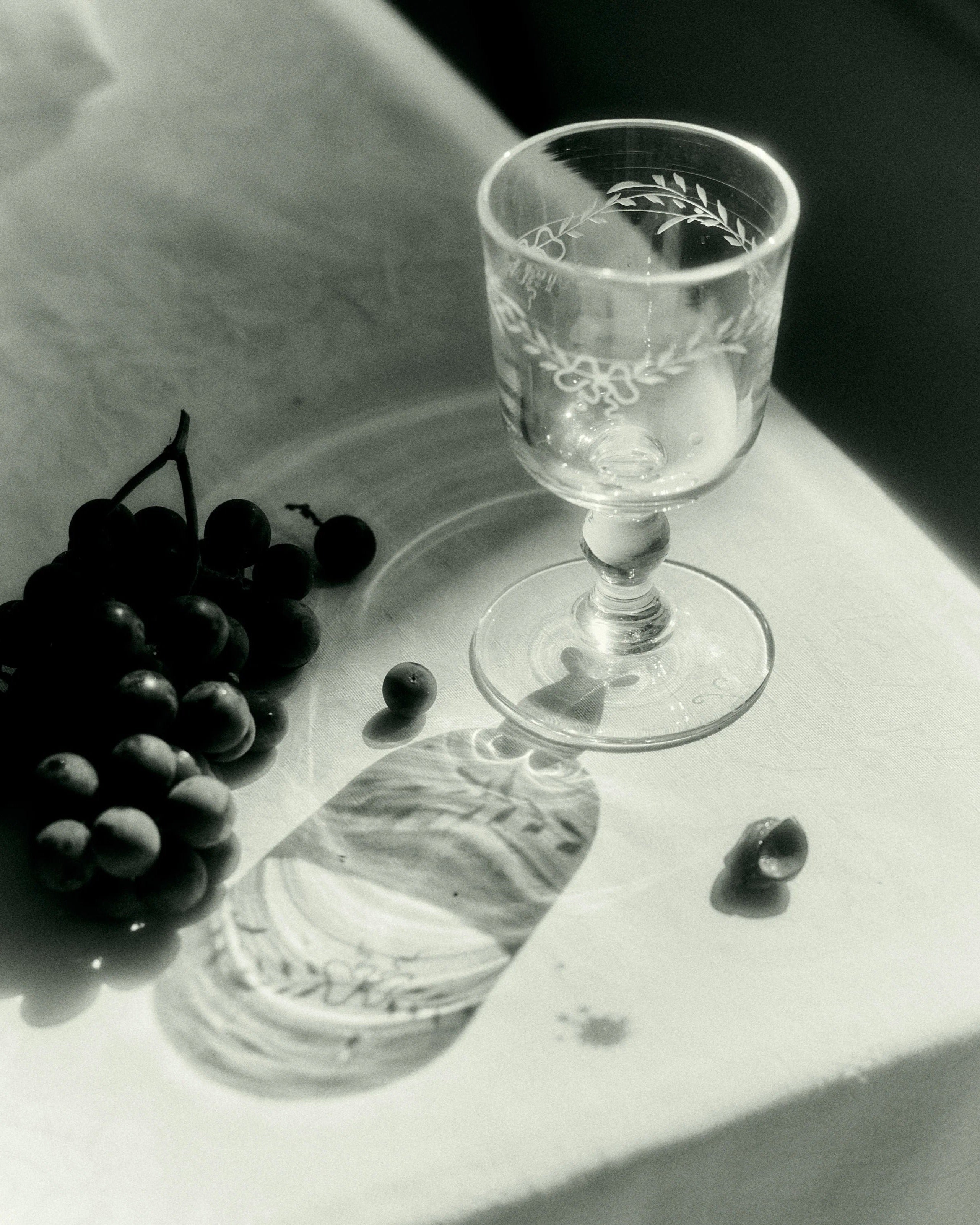 Black and white photo of a cluster of grapes next to an ornate Akua Objects Barbro Wine Glass - Set of four on a table, with light casting a detailed shadow of the glass.
